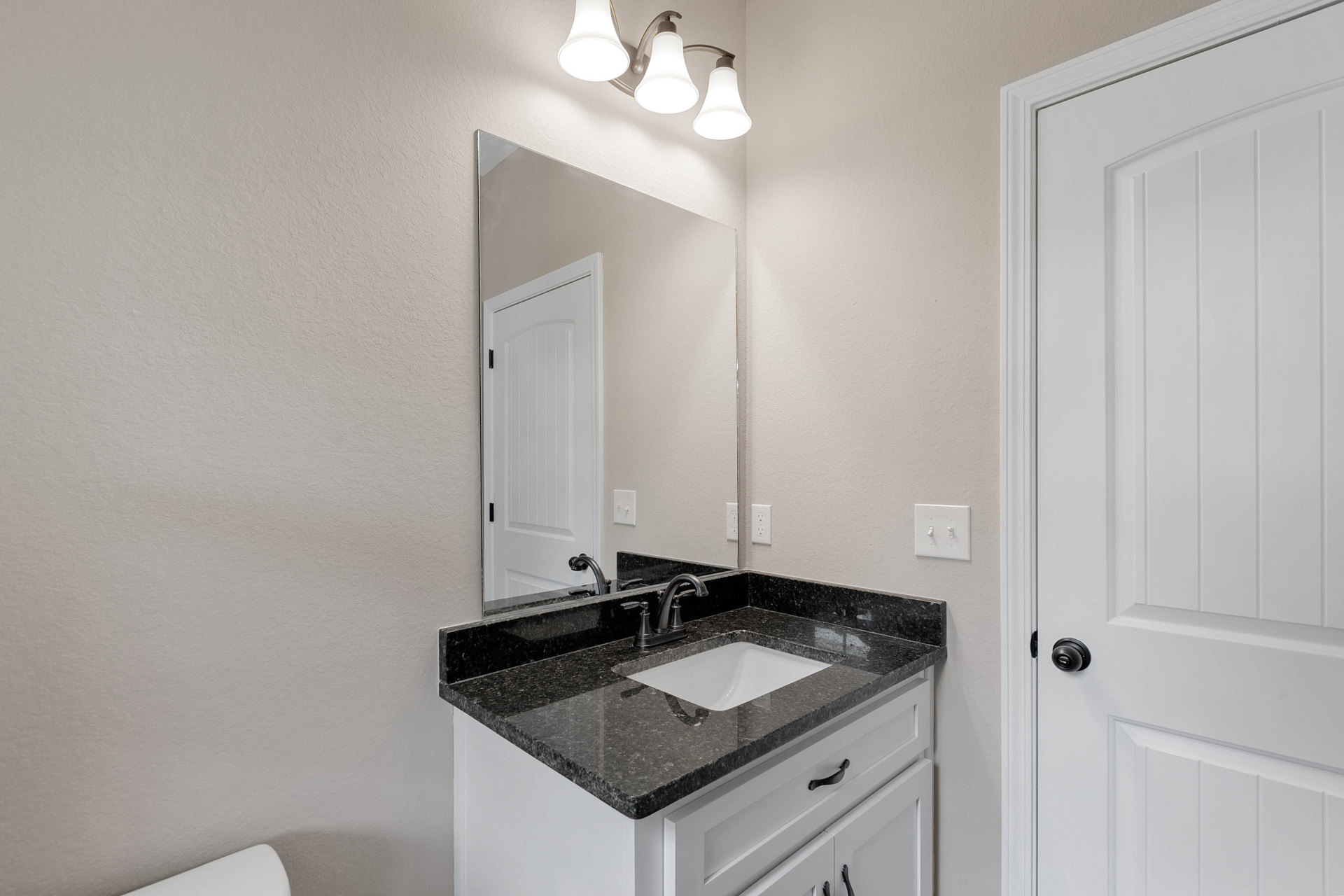 Bathroom with black stone countertop, rectangular mirror above sink, white paneled door, white bathtub, chrome faucet, tile walls, and modern light fixture.