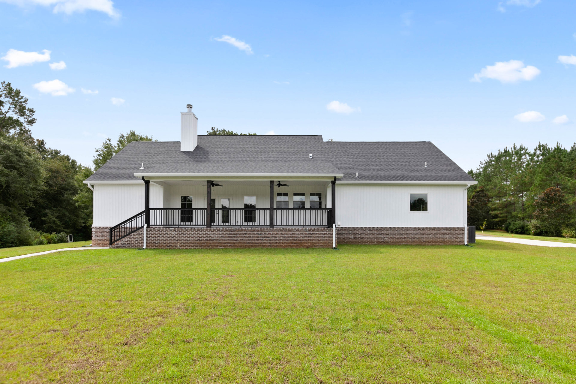 White siding farmhouse with black porch railing, manicured green lawn, brick retaining wall, and mature trees under a blue sky