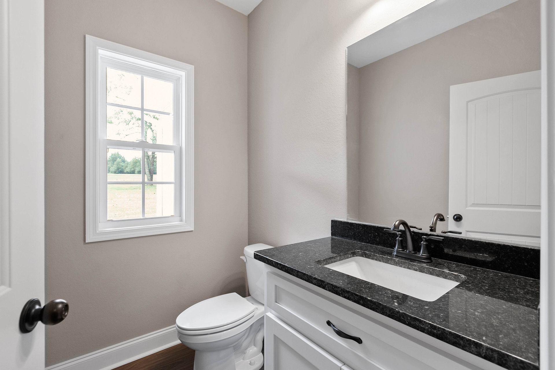 Bathroom with black stone countertop, white toilet, rectangular window showing leafy tree, white paneled door, freestanding white bathtub, chrome faucet and sink, light tile