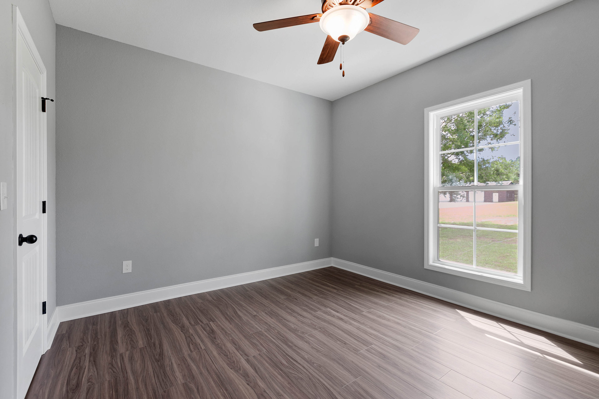 Ceiling fan with integrated light fixture, large window overlooking trees, wood laminate flooring, white door with black handle, smooth plaster walls