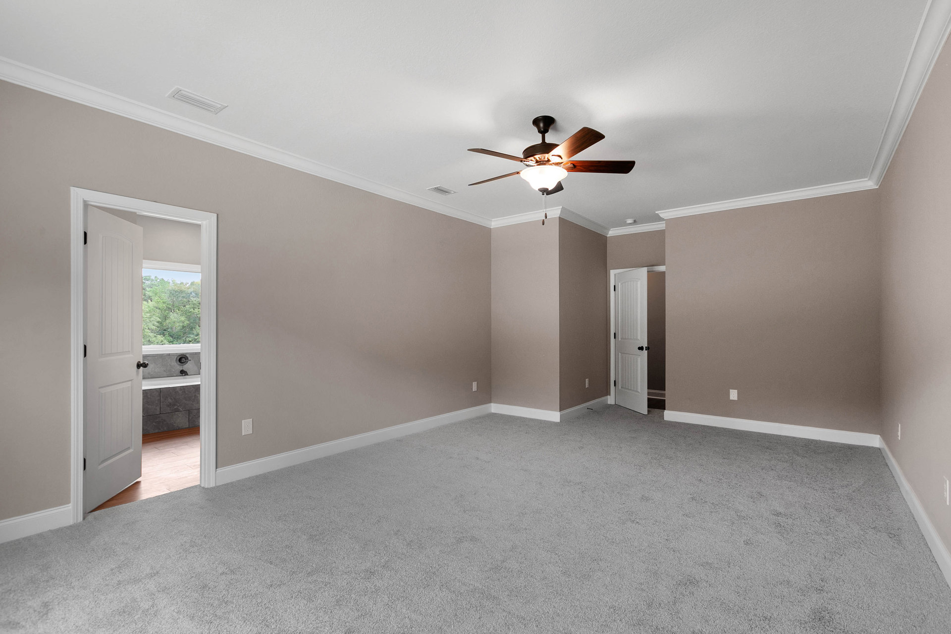Ceiling fan with light fixture above white carpeted floor, open door leading to bathtub, white door with black knobs, large window offering forest view.