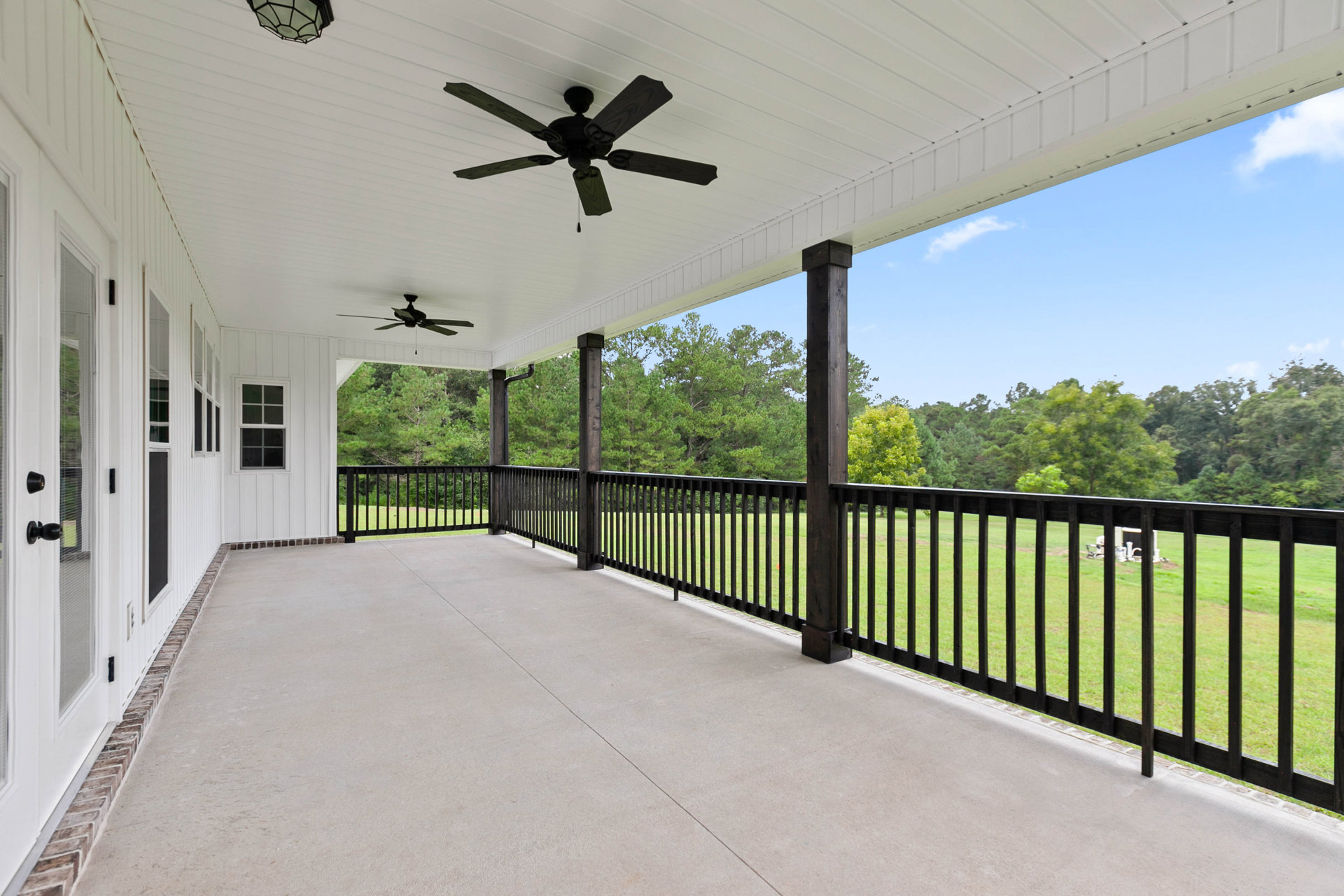Porch with black iron railing, black-bladed ceiling fan, concrete deck, wooden support post, and fenced grassy yard