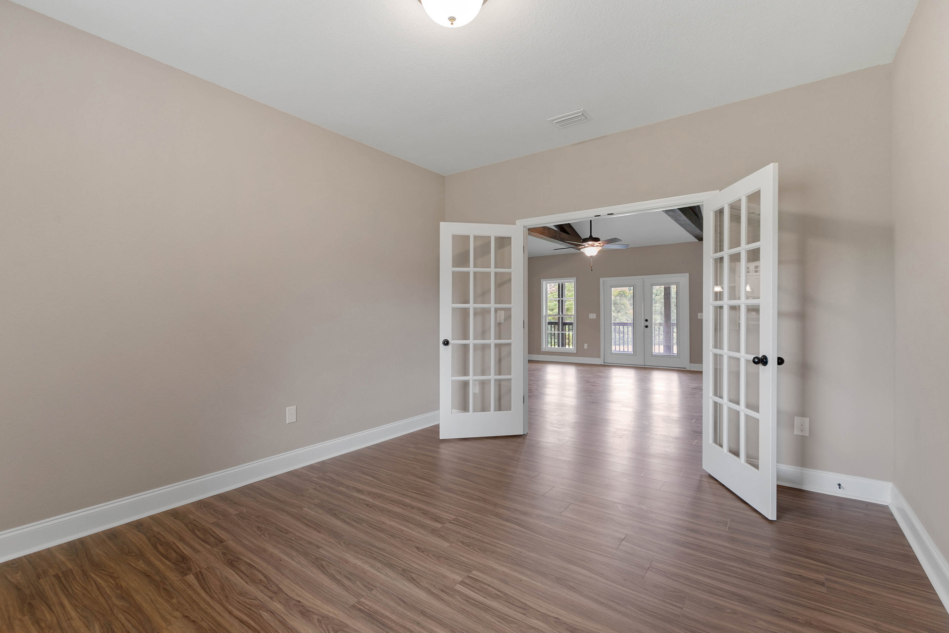 Wood flooring in a bright room with white double doors featuring glass panes, white walls, and a window overlooking trees.