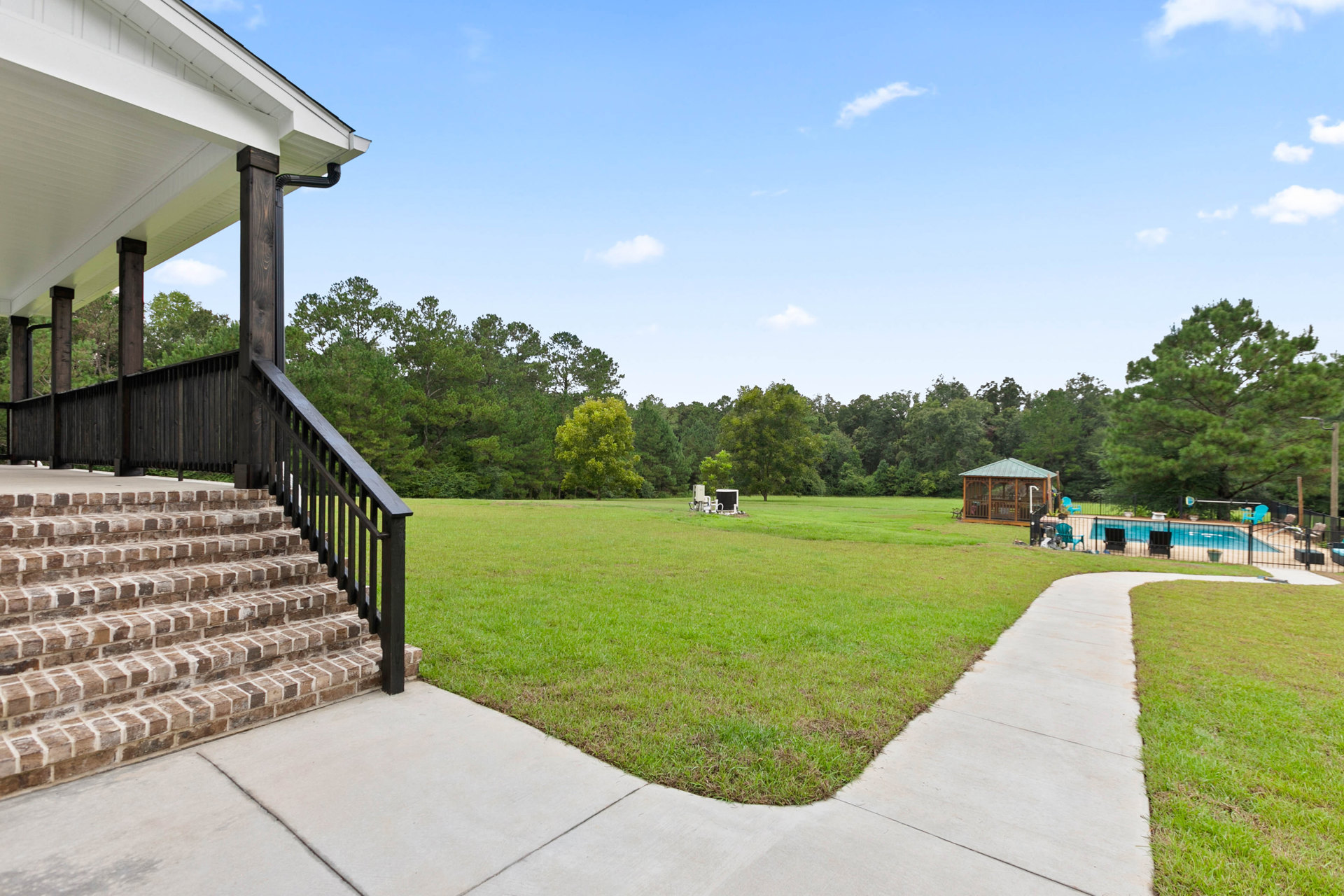 Two-story house with white siding, black roof, and large windows, surrounded by a green lawn; wooden gazebo with green roof in backyard; black metal railings on stairs; mature tree