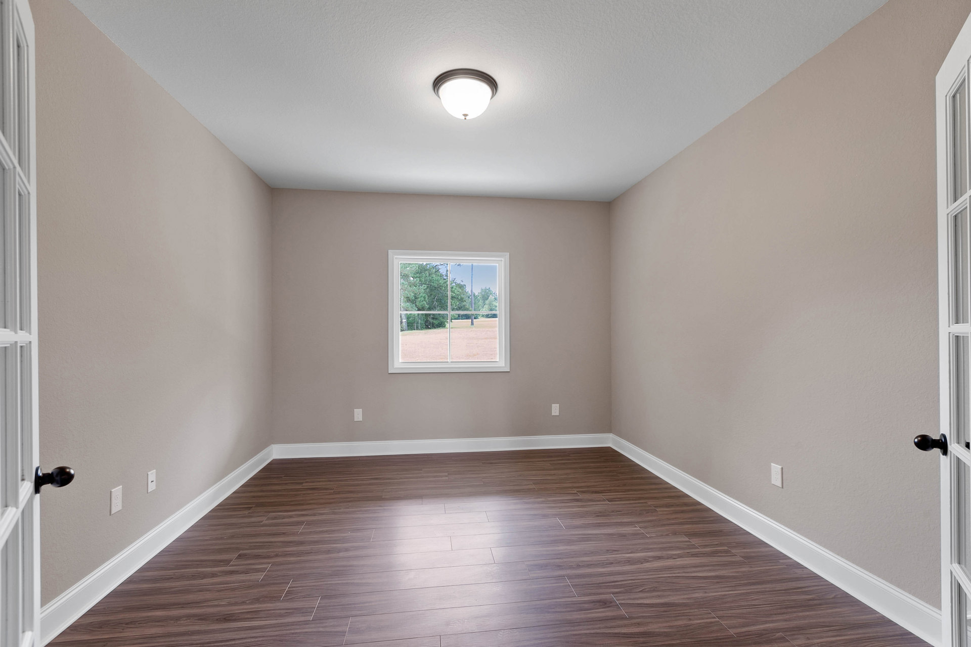 Sunlit room featuring wide-plank hardwood flooring, white baseboard trim, large window overlooking leafy trees, smooth plaster walls, and a modern brushed metal light fixture.
