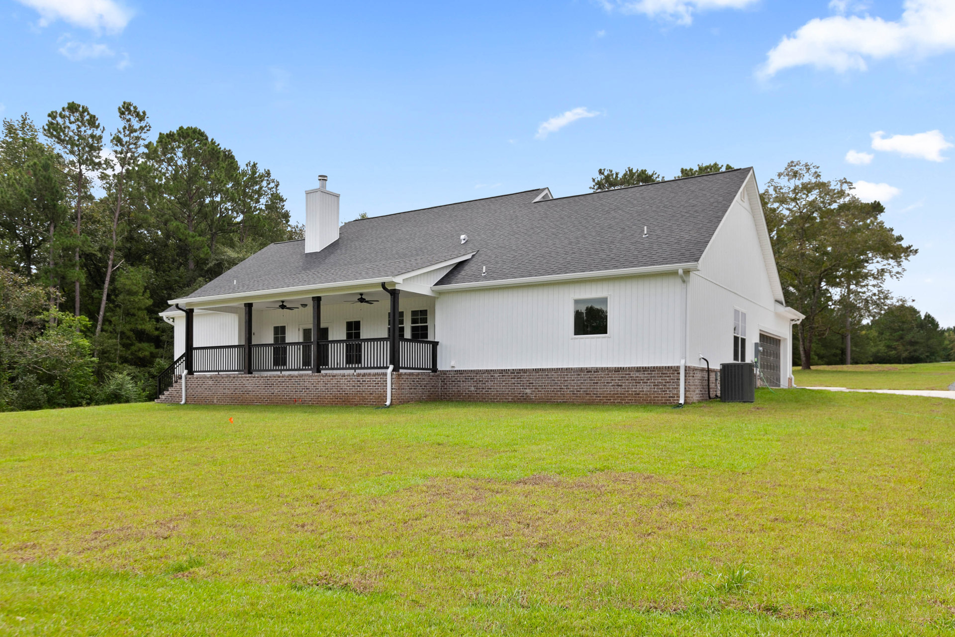 White farmhouse with black porch railing, brick foundation, large windows, manicured lawn, and mature trees in the background