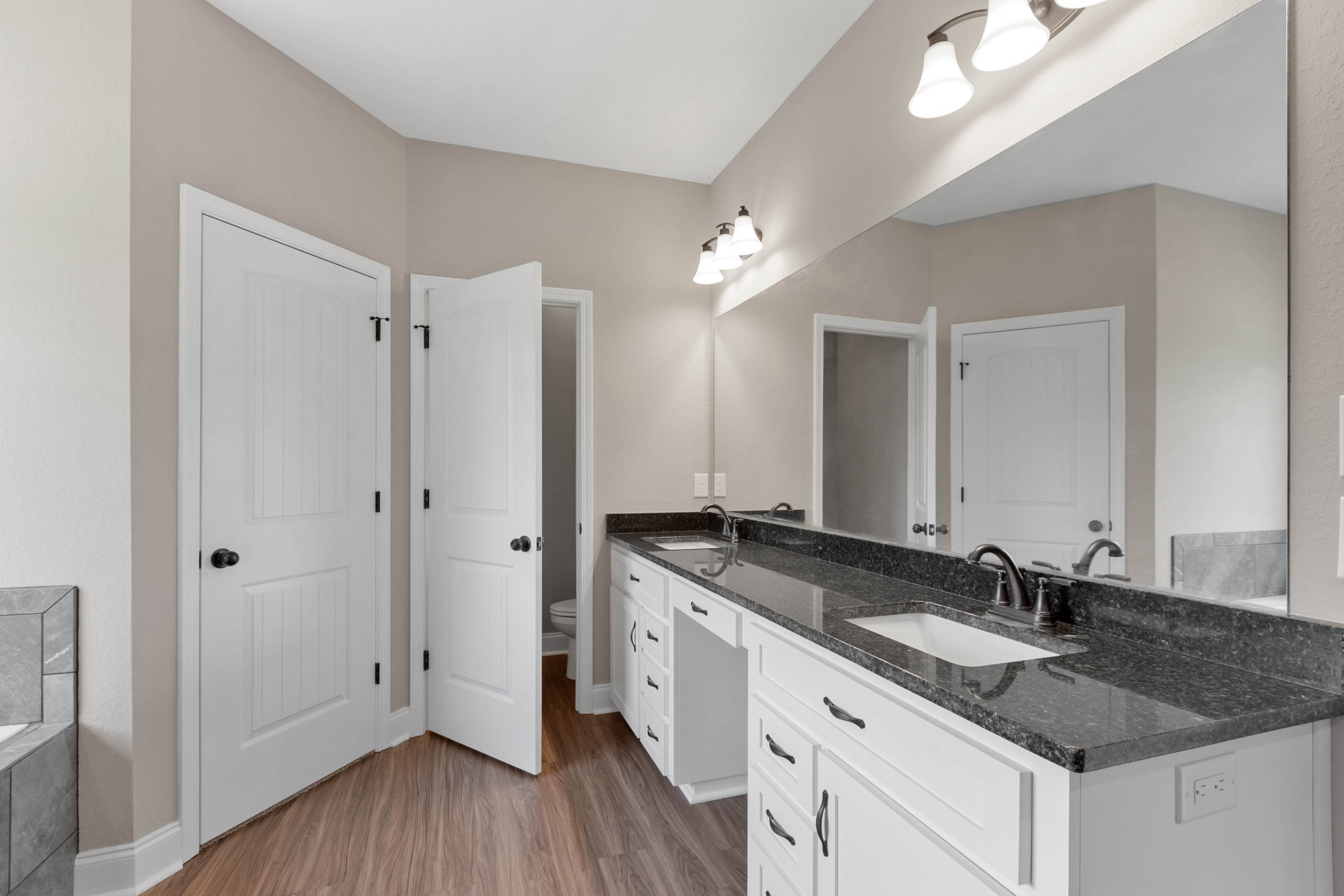 Bathroom featuring white shaker cabinets, matte black countertops, undermount sink with chrome faucet, large frameless mirror, white door with black handle, and neutral tile
