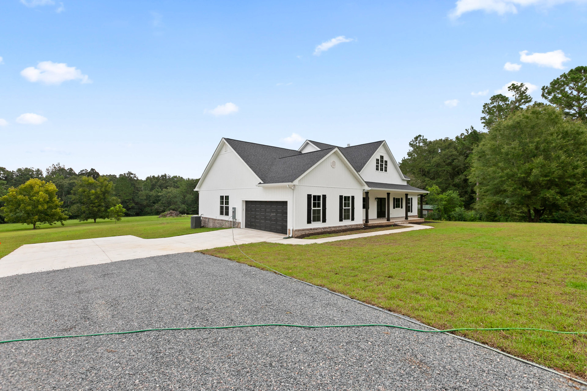 White two-story home with black garage doors, concrete driveway, green lawn, and mature tree in front yard under partly cloudy sky