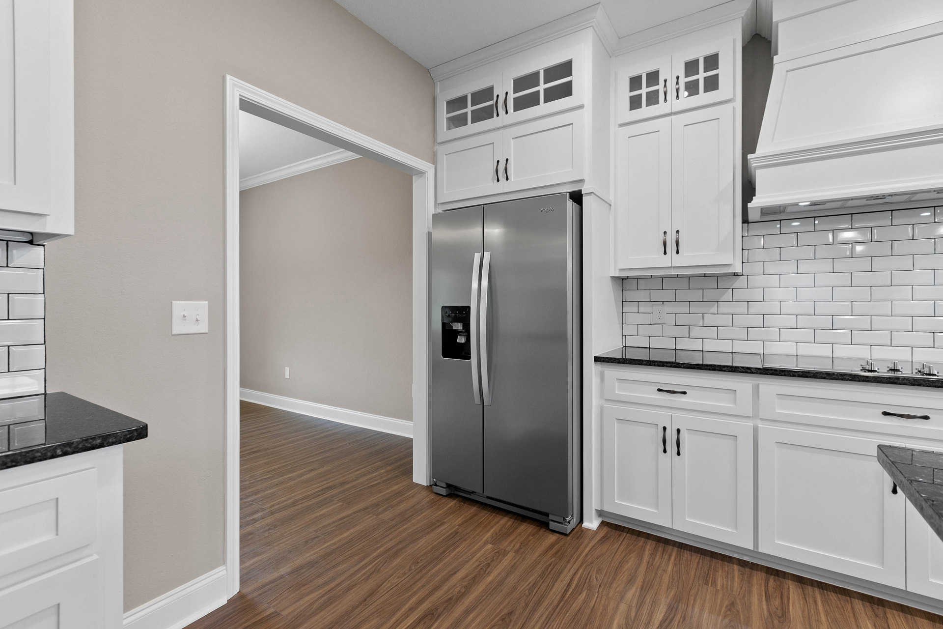 White kitchen with shaker cabinets, black countertops, stainless steel refrigerator with double handles, white range hood above stove, wood flooring with white baseboards, light