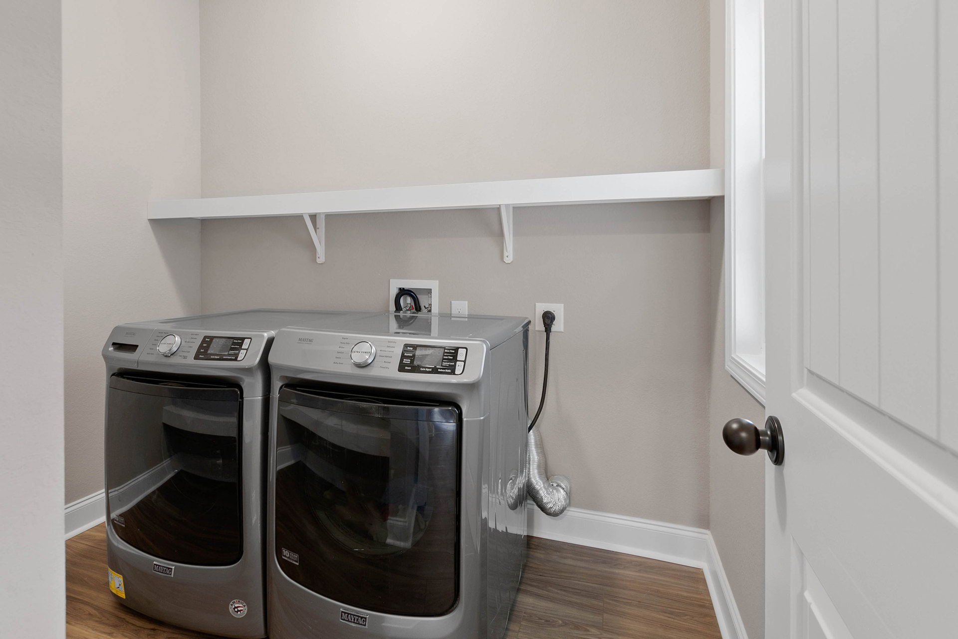 Front-loading washing machine with white control knob, black cable emerging from wall, light-colored cabinetry and countertop, tiled floor, neutral painted walls