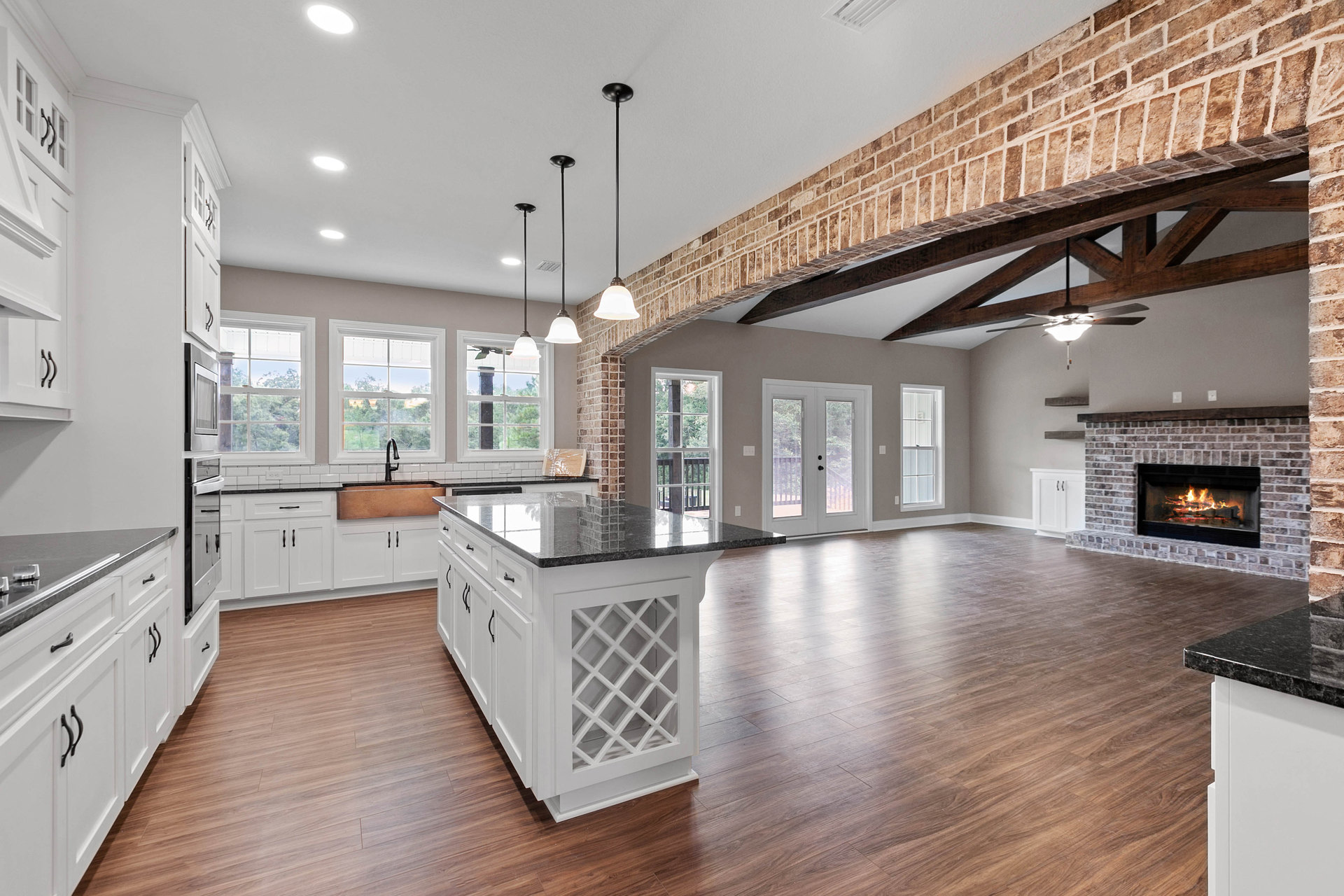 Spacious open floor plan featuring a brick fireplace, large kitchen with marble-topped island, white cabinetry, stainless steel appliances, and a window above the sink overlooking