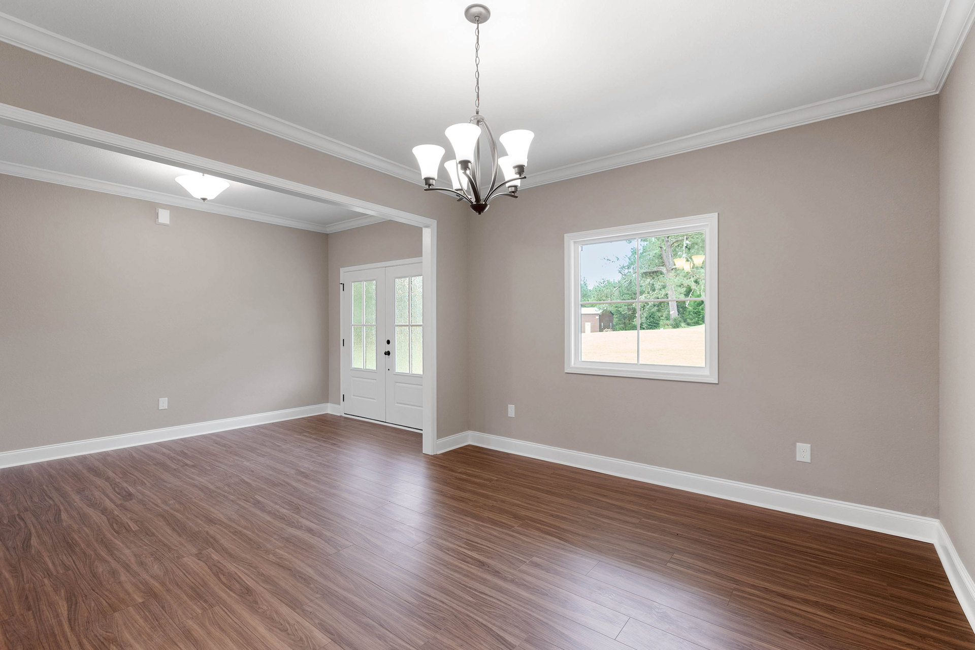 Wood flooring in a spacious room with a ceiling-mounted chandelier, double doors with frosted glass, and a large window overlooking a tree and neighboring house.