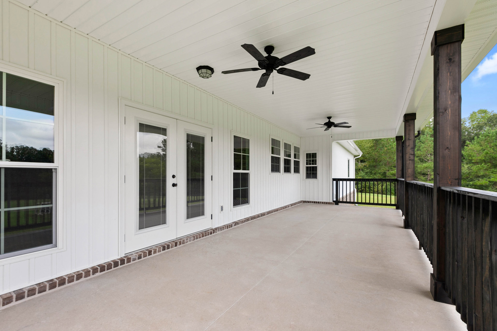 White porch with black metal railing, four-blade black ceiling fan, concrete patio, white-framed windows reflecting a fence, and glass door.