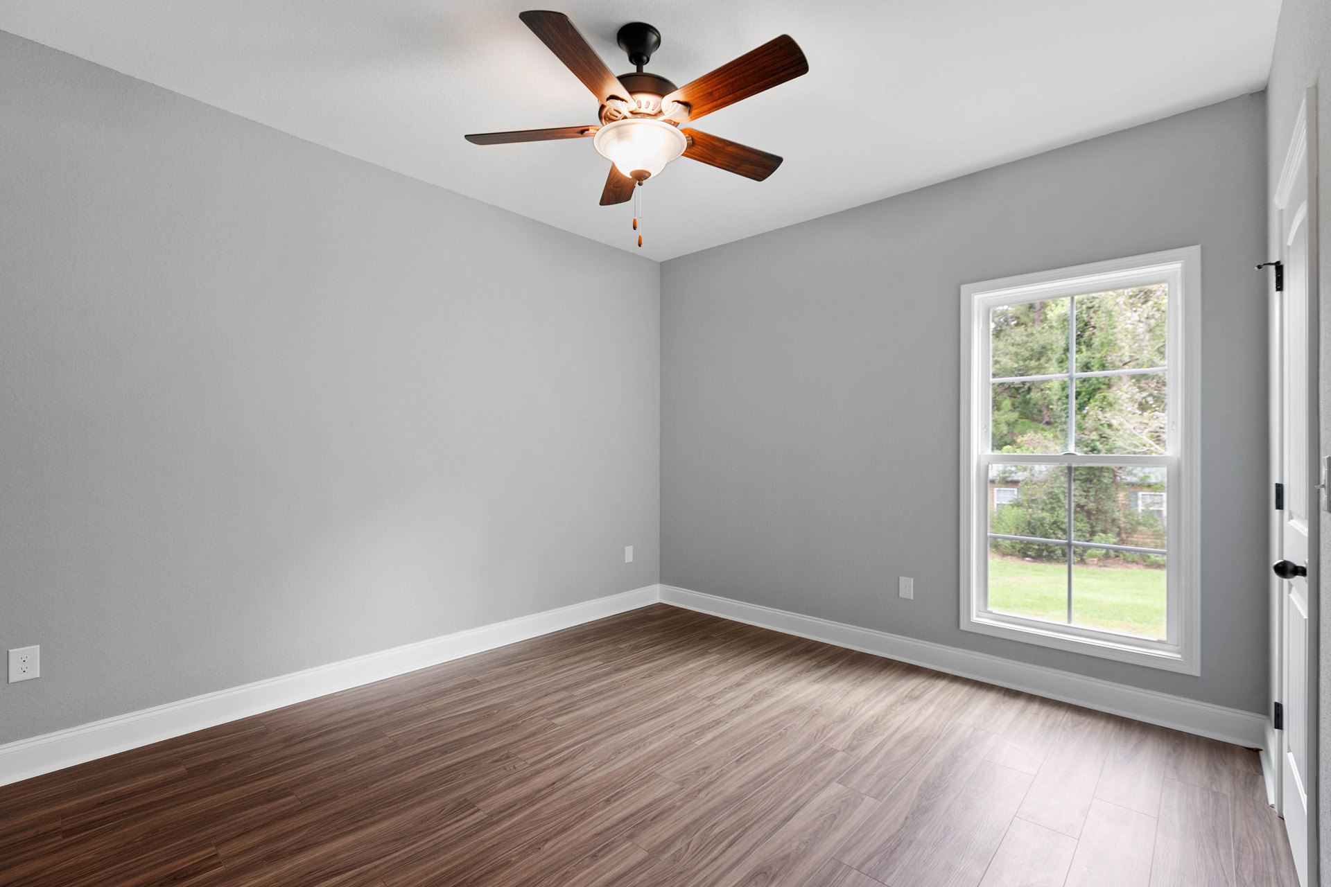 Ceiling fan with integrated light fixture mounted above wood flooring and white baseboards, window revealing trees outside