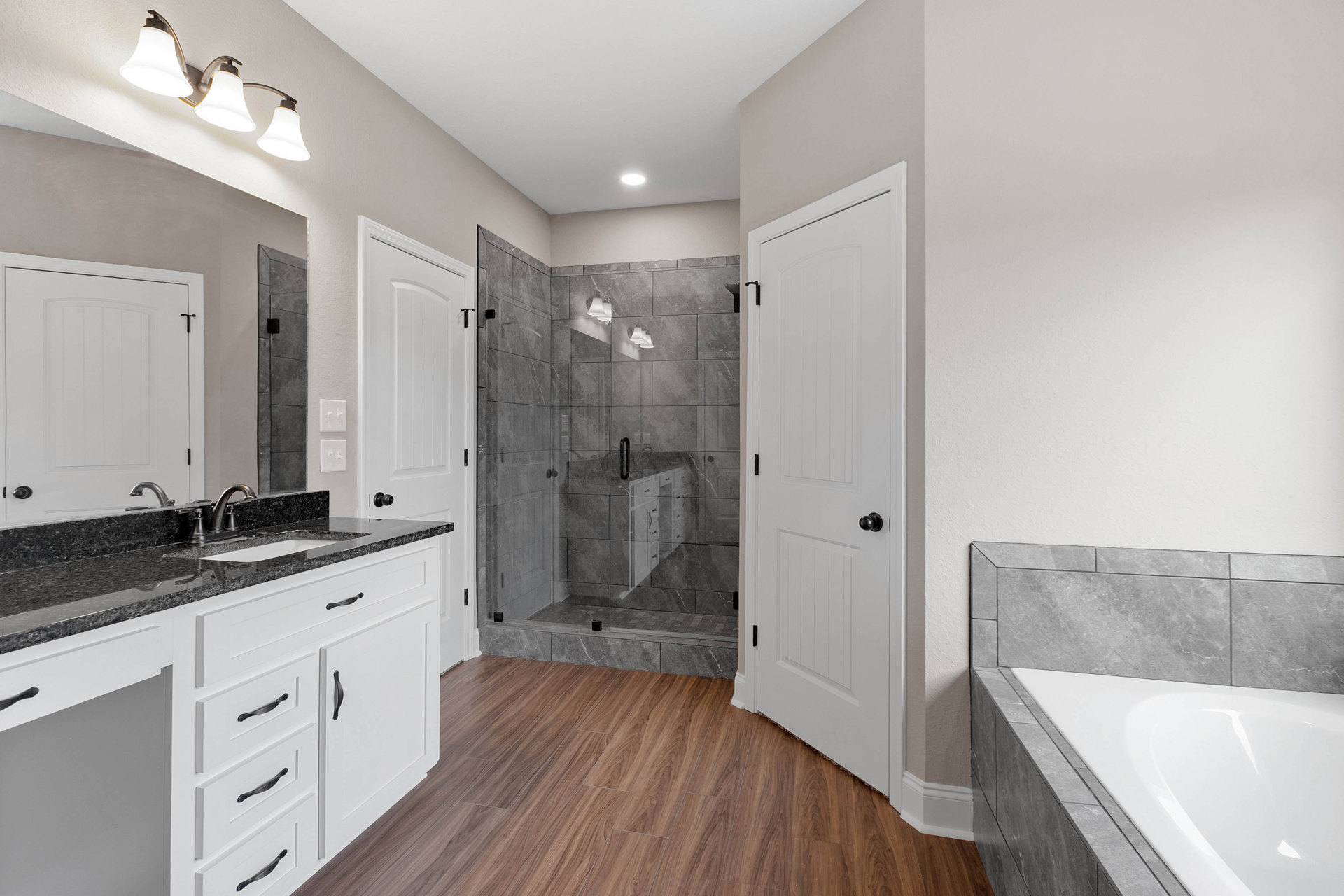 Bathroom featuring a frameless glass shower, freestanding white bathtub with grey tile surround, wood flooring, stone ledge, white cabinetry, black door knobs, and a three-lamp