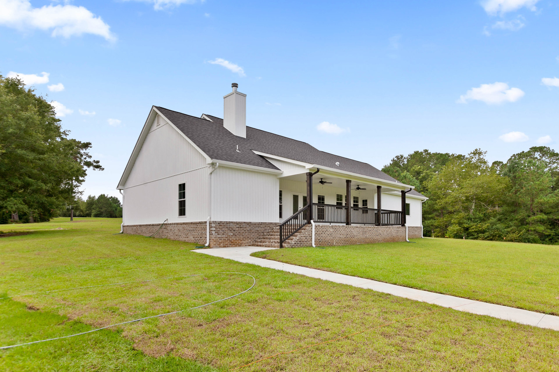 Two-story farmhouse with gray siding, white trim, covered front porch, brick chimney, manicured lawn, stone walkway, leafy tree, blue sky with clouds