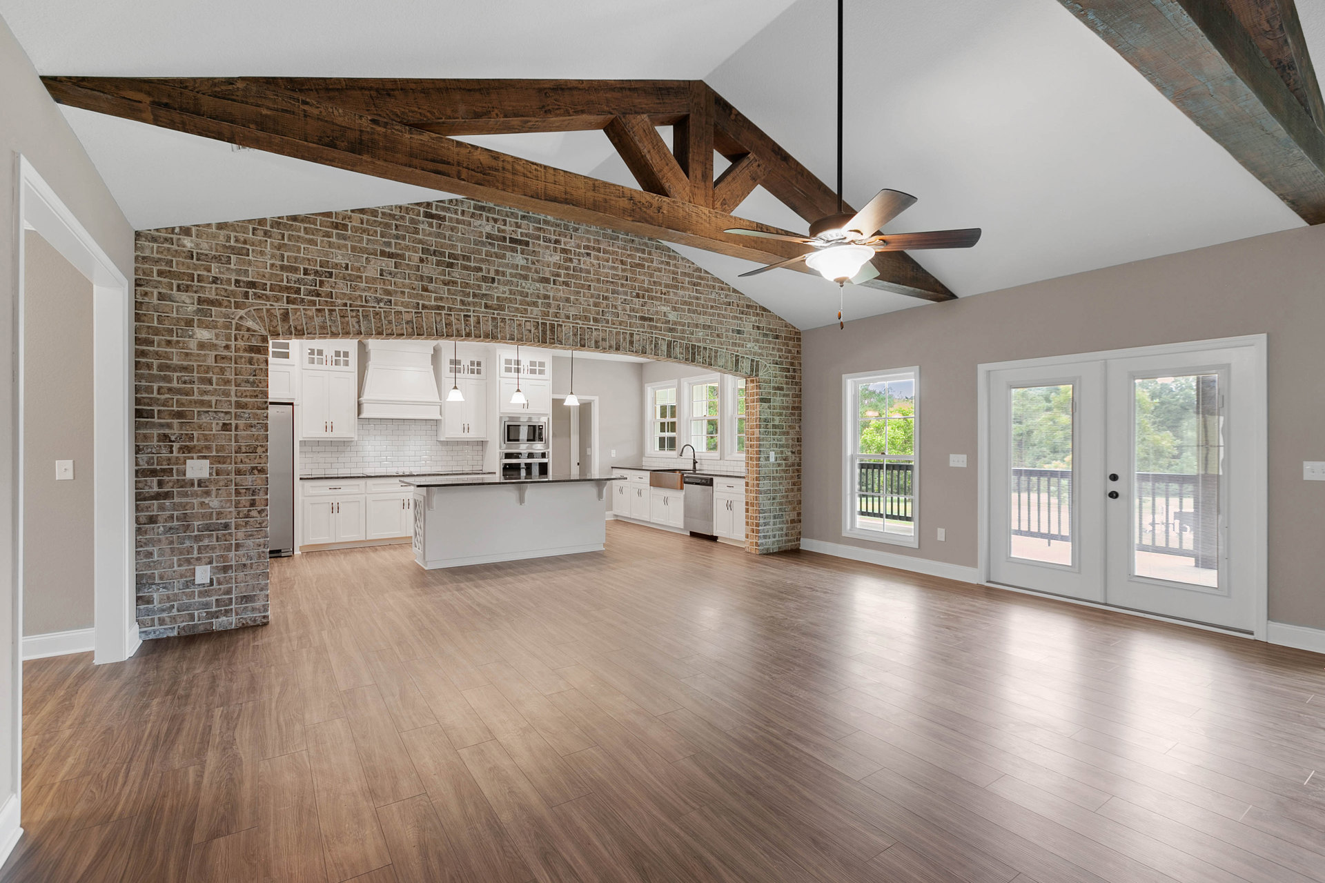 Spacious open floor plan featuring a prominent brick wall, wood flooring, ceiling fan, white kitchen cabinetry with black countertops, brick column, fireplace, double glass-paneled
