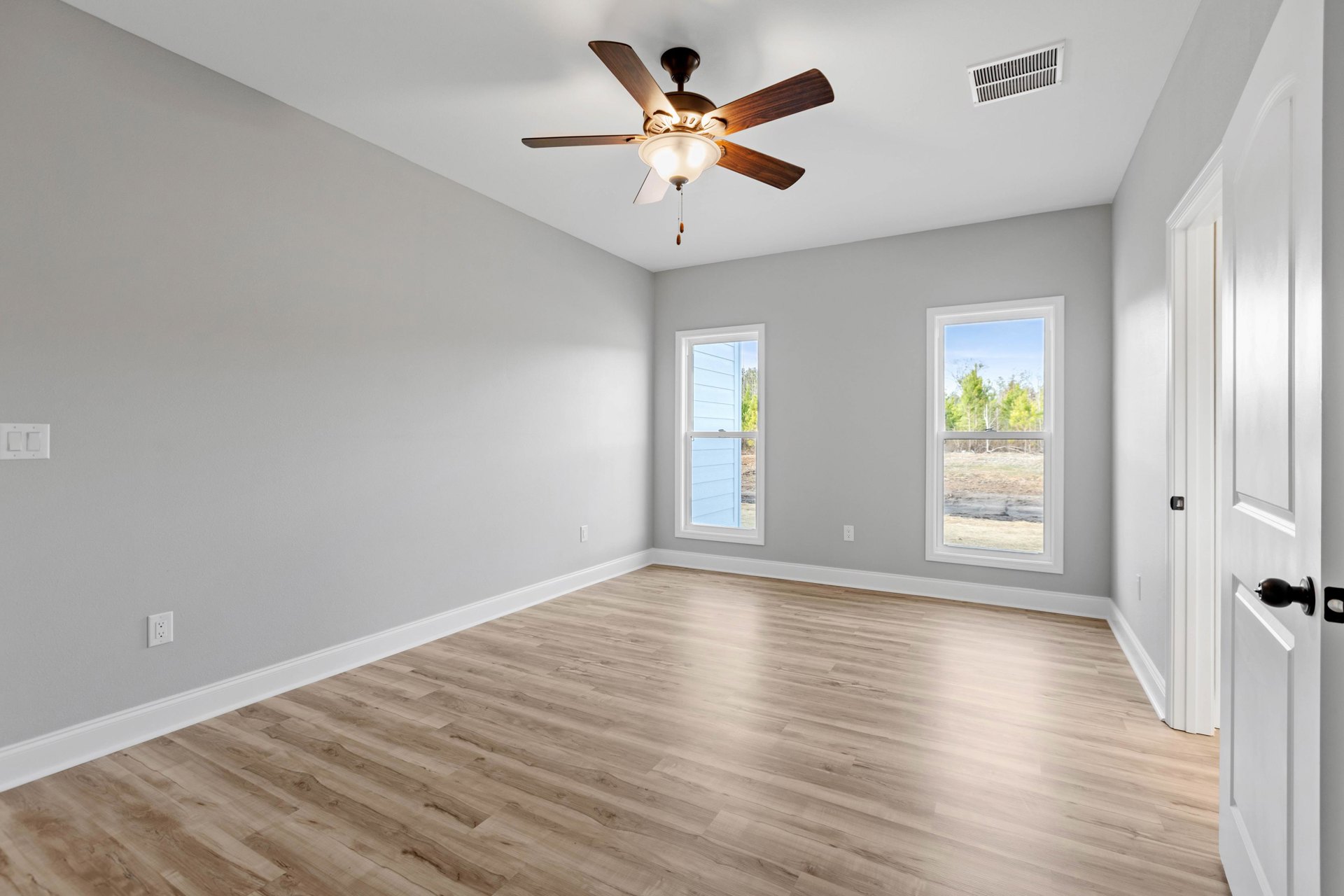 Ceiling fan with light fixture, two large windows with white frames overlooking trees, hardwood flooring, white walls, and a white light switch