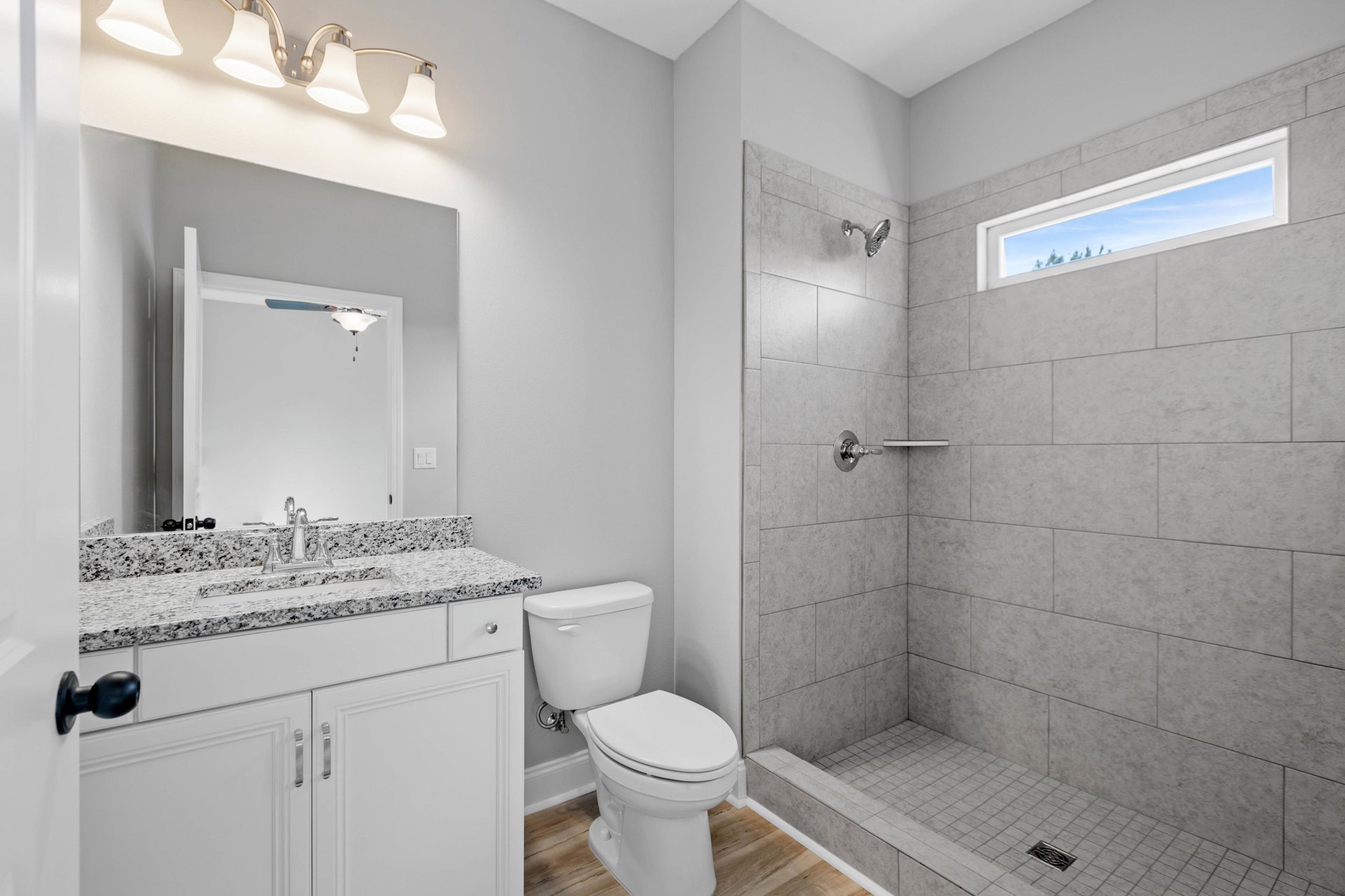 White toilet in corner beside glass shower enclosure, window with blue sky above, dual bulb light fixture mounted on wall, neutral tile flooring and walls