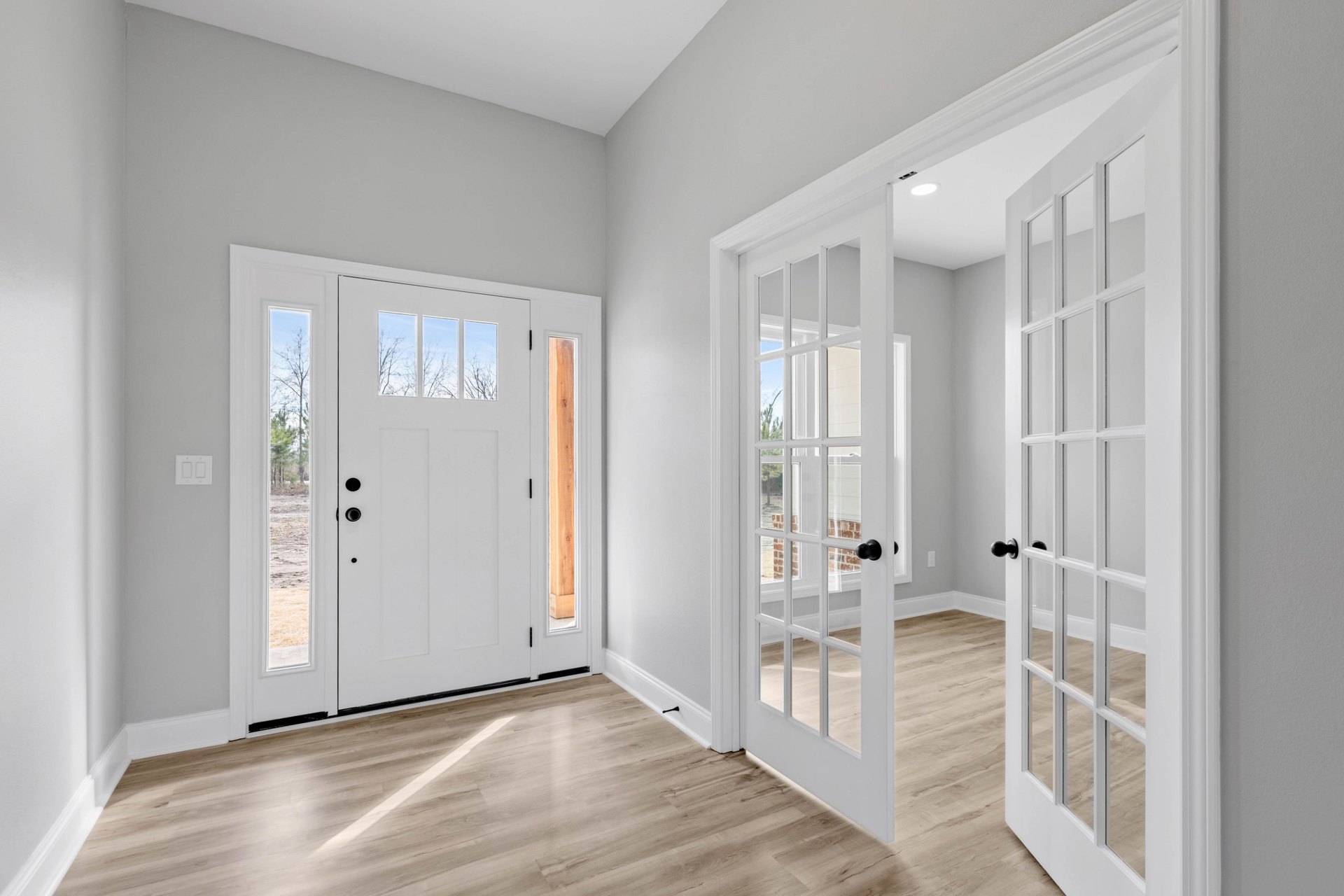 White walls and doors, wood flooring with visible black screw in corner, window showing trees outside
