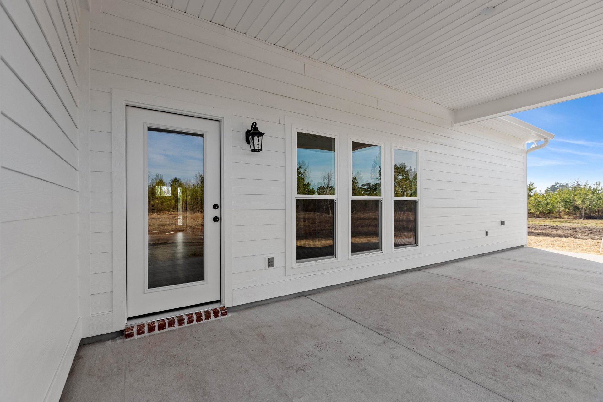 White house with white walls and door, black light fixture mounted beside entry, concrete porch floor, glass door offering view of forest in background.