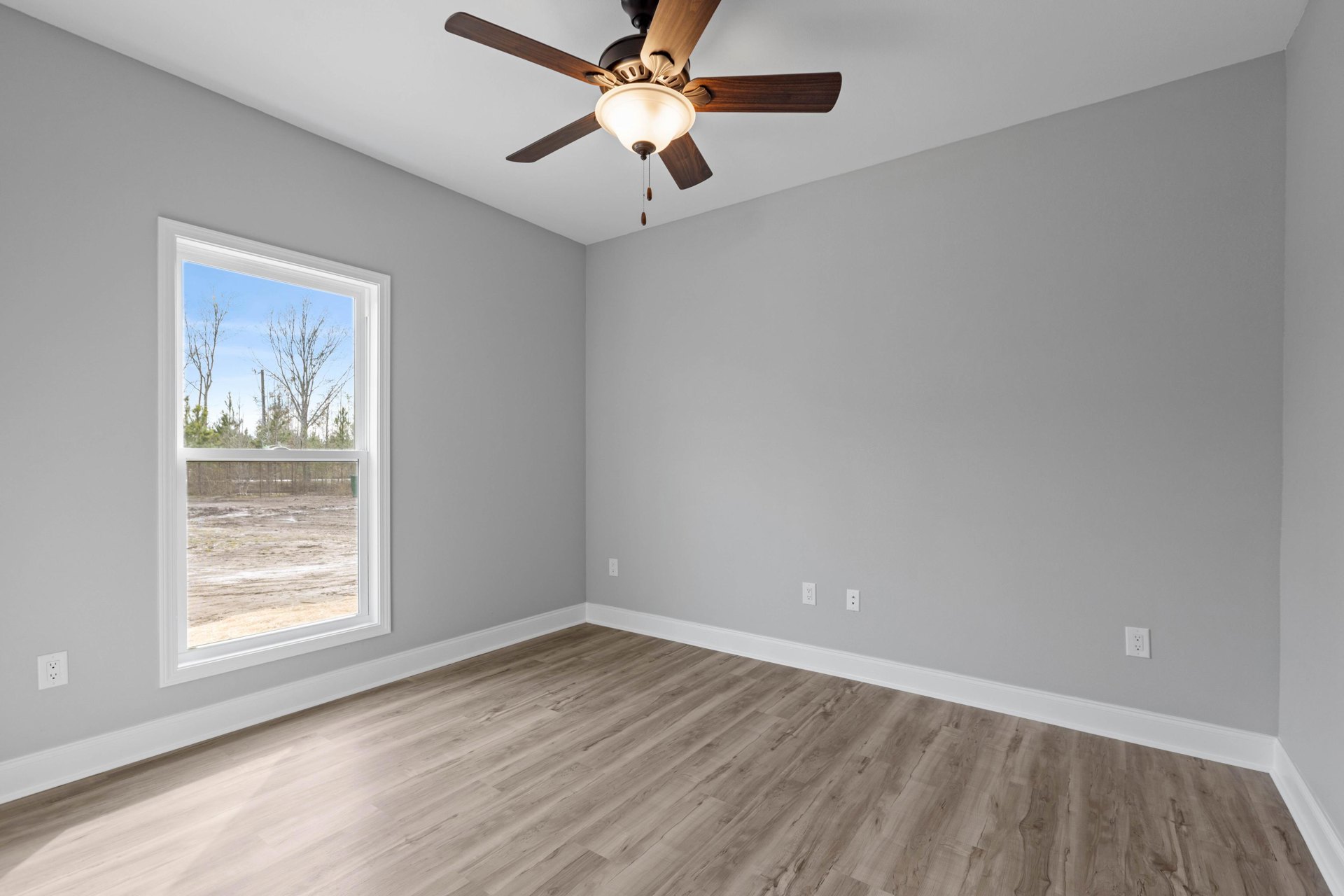 Ceiling fan with integrated light fixture mounted on white plaster ceiling, wood flooring with white trim, large window overlooking dirt field and trees.