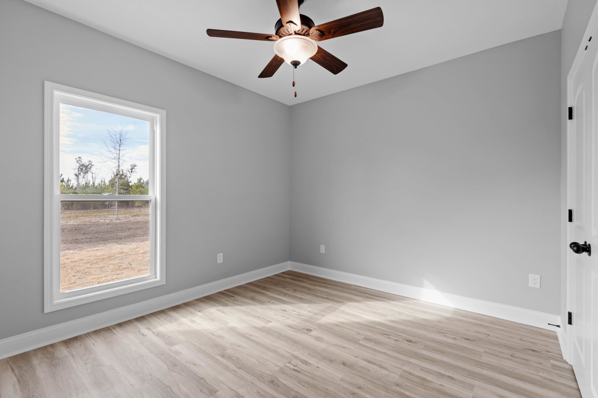 Wood flooring and white walls in a room featuring a ceiling fan with light fixture, large window overlooking dirt field and trees, and a close-up of a door knob.