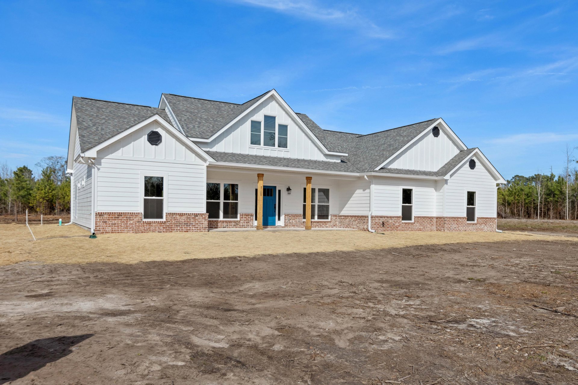 White clapboard house with blue door and screened window, dirt road leading past a dirt field toward a brick building resembling Robert Frost Farm, tree and cloud-filled sky in