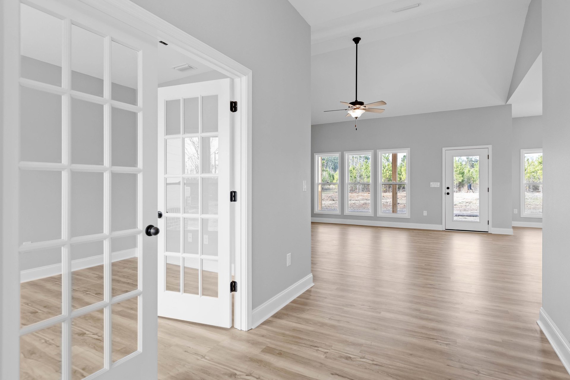 White-walled room with wood laminate flooring, ceiling fan with light fixture, and white door featuring glass panes; outdoor trash can visible through door.
