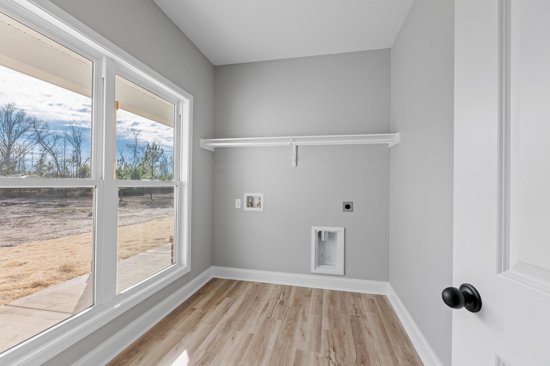 Wood floor with white trim, white shelving, window overlooking dirt field and trees, white refrigerator with silver cylinder, black round door knob
