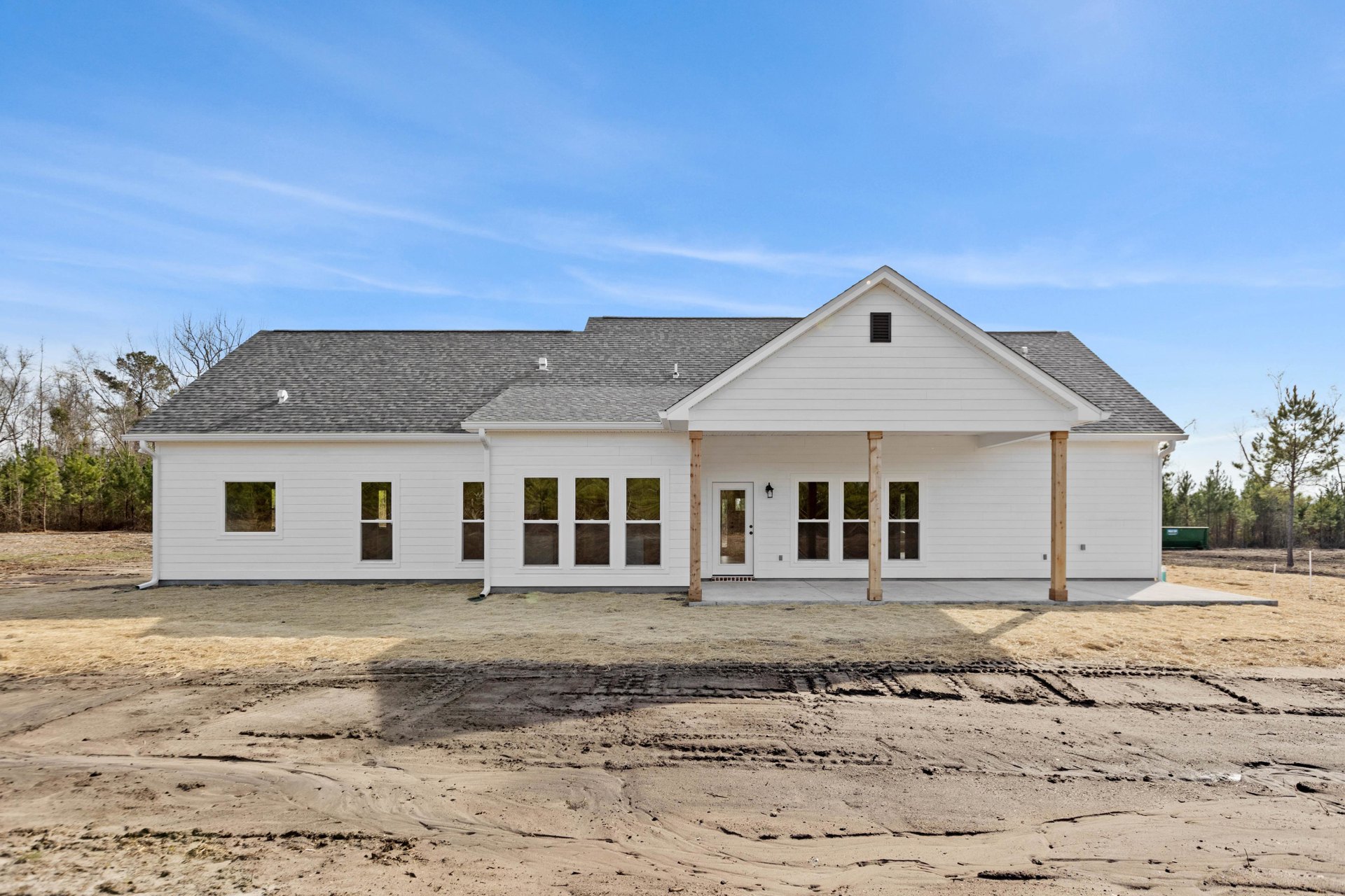White siding house with wide covered porch, gray shingle roof, multiple windows, large driveway, surrounded by dirt and grass field under blue sky with scattered clouds.