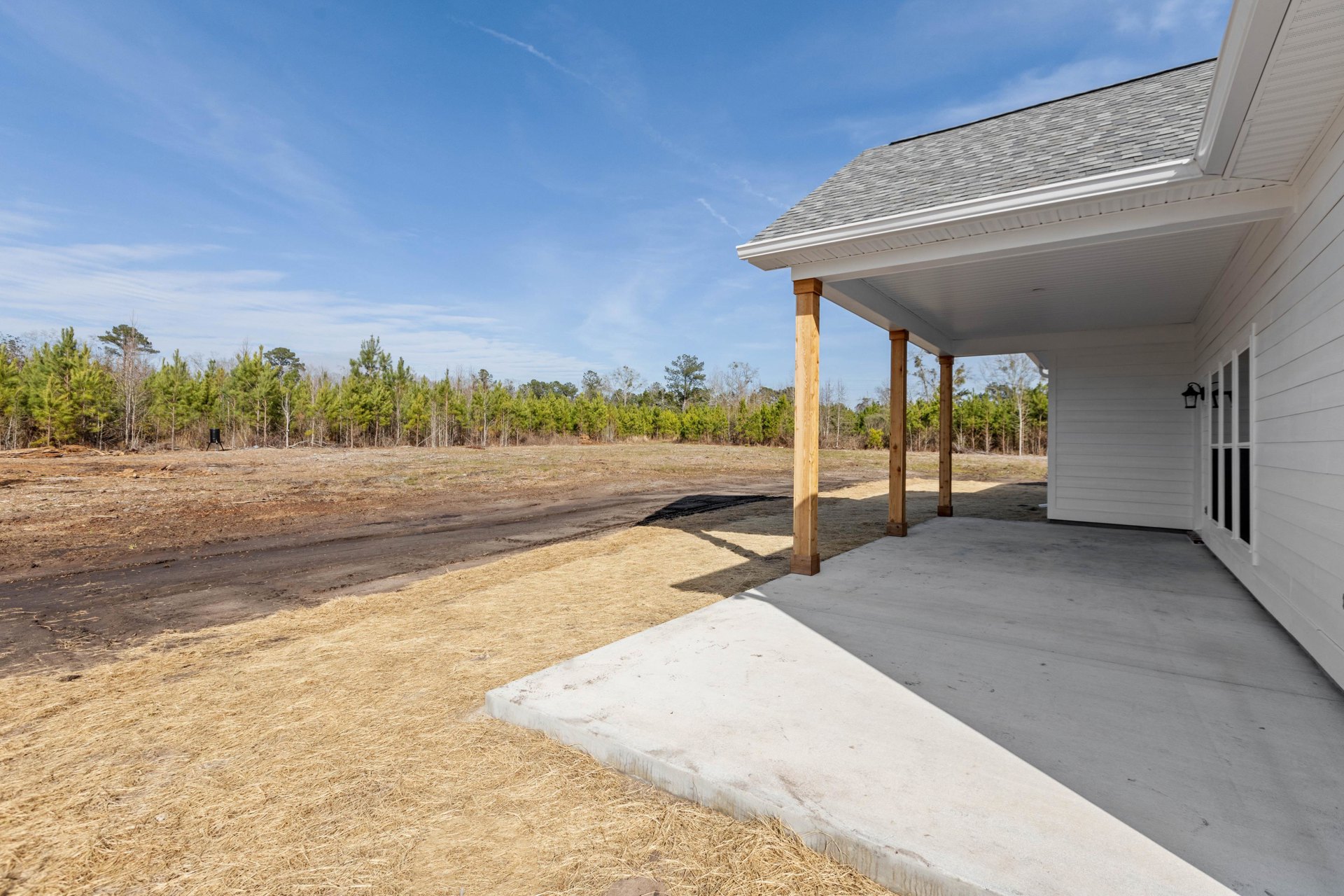 Covered porch with wooden pillars and concrete slab, light-colored exterior walls, dry grass and dirt road in foreground, blue sky with scattered clouds above
