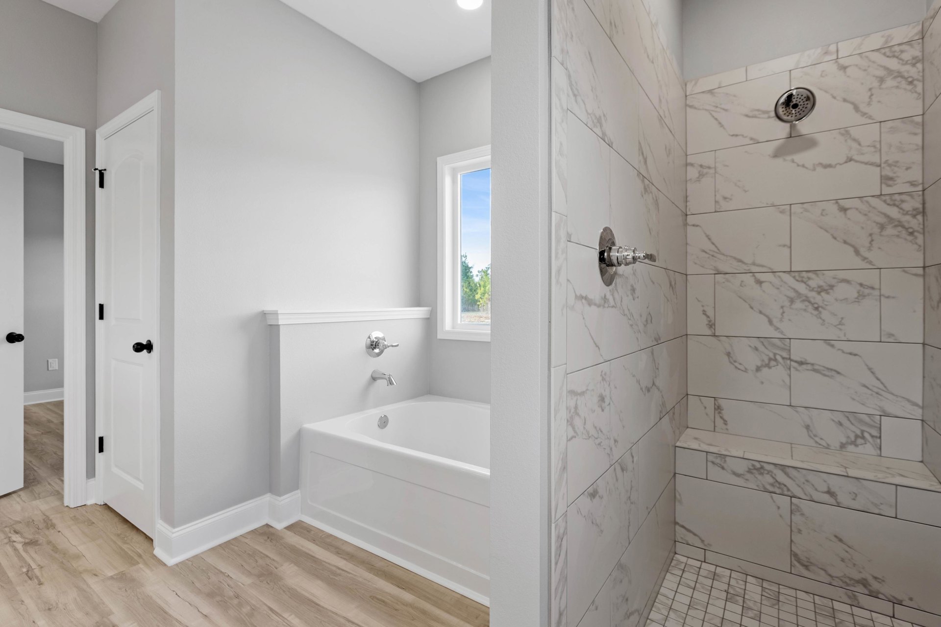 White tile bathroom featuring a freestanding bathtub, glass-enclosed shower with black and white marble accents, chrome shower head and faucet, window overlooking trees and blue