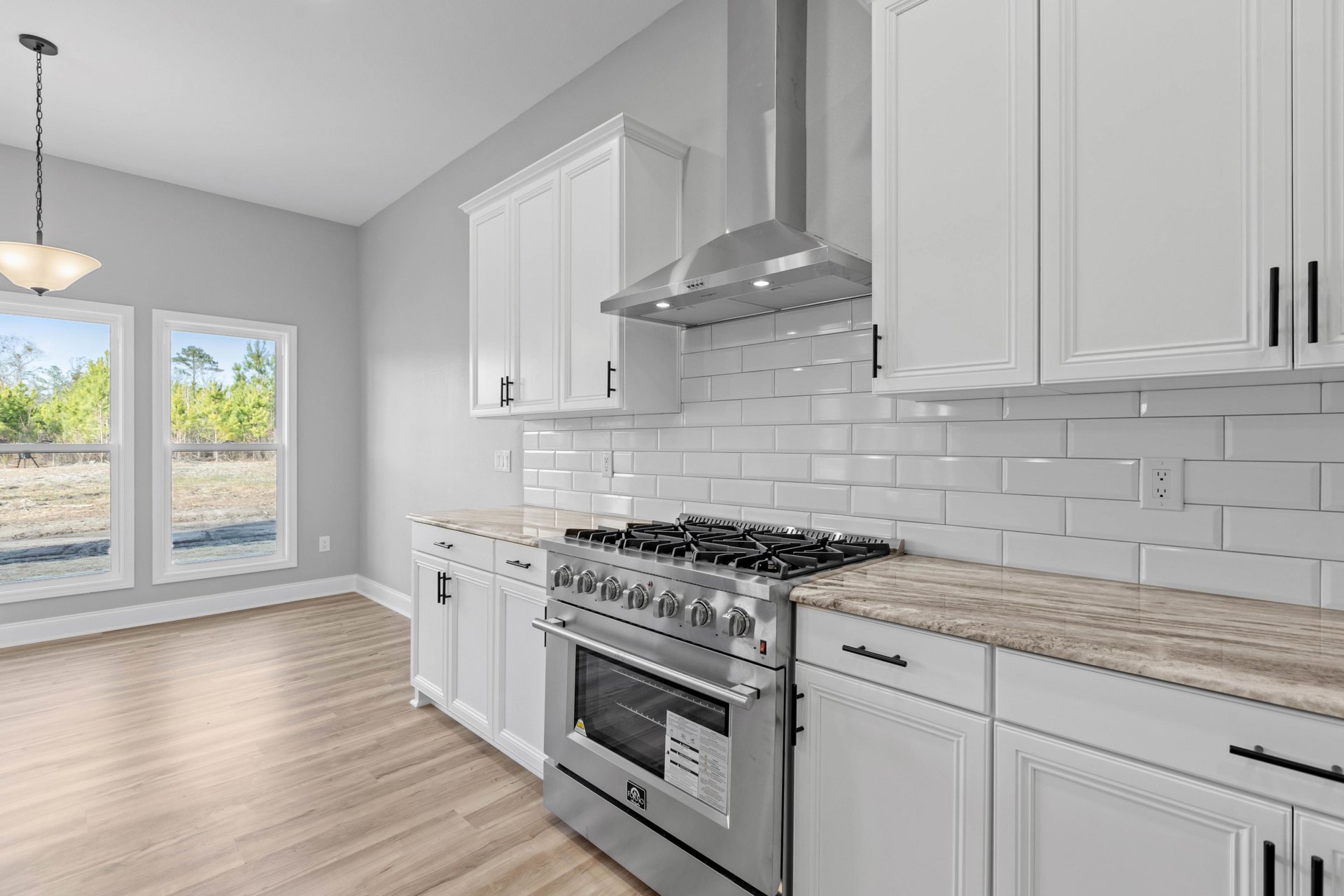 White kitchen cabinets, stainless steel stove with glass oven door, stone countertop, window overlooking trees.