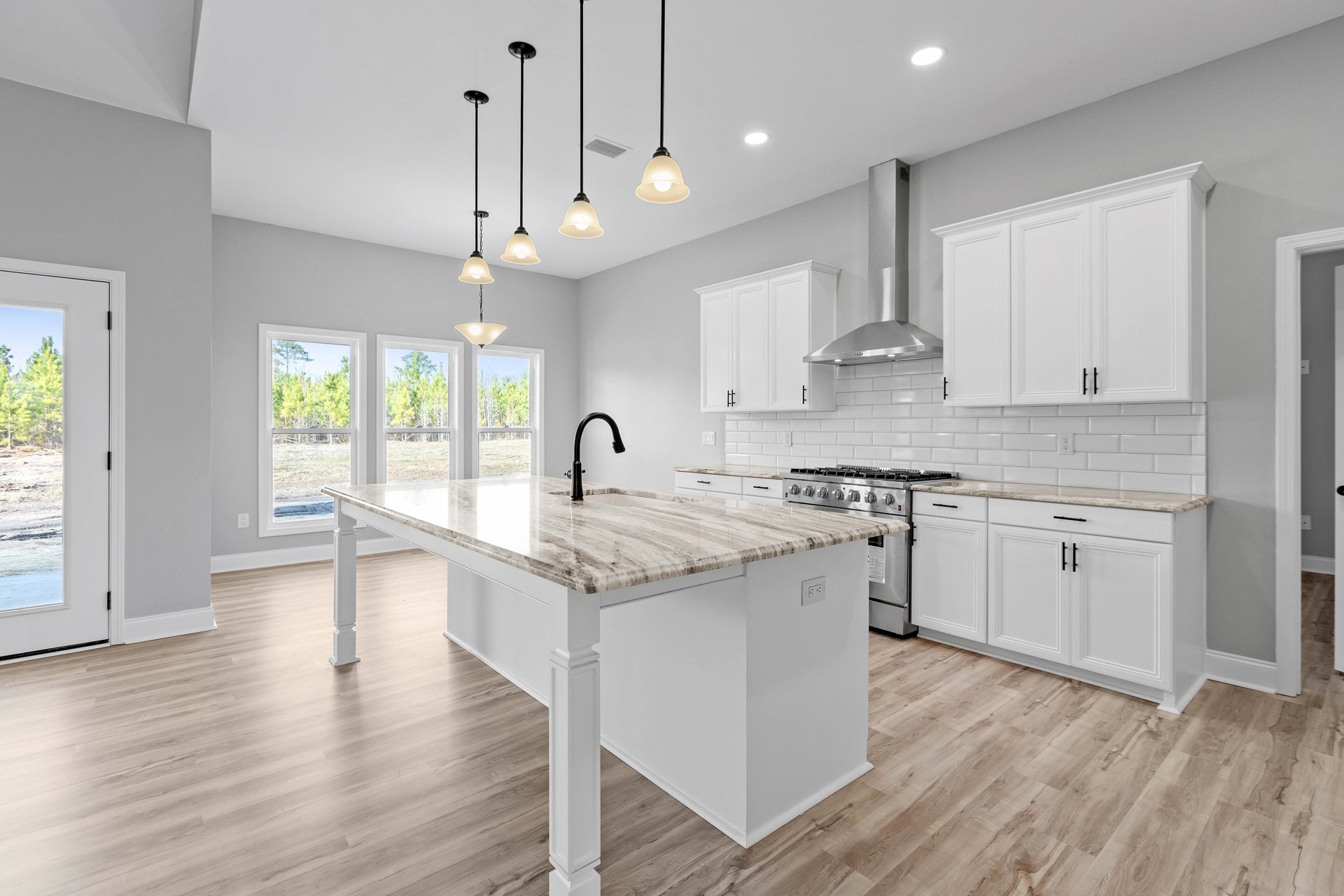 Kitchen with white cabinets and black handles, central island featuring a black faucet, stove, tile backsplash, pendant light, and window overlooking trees.