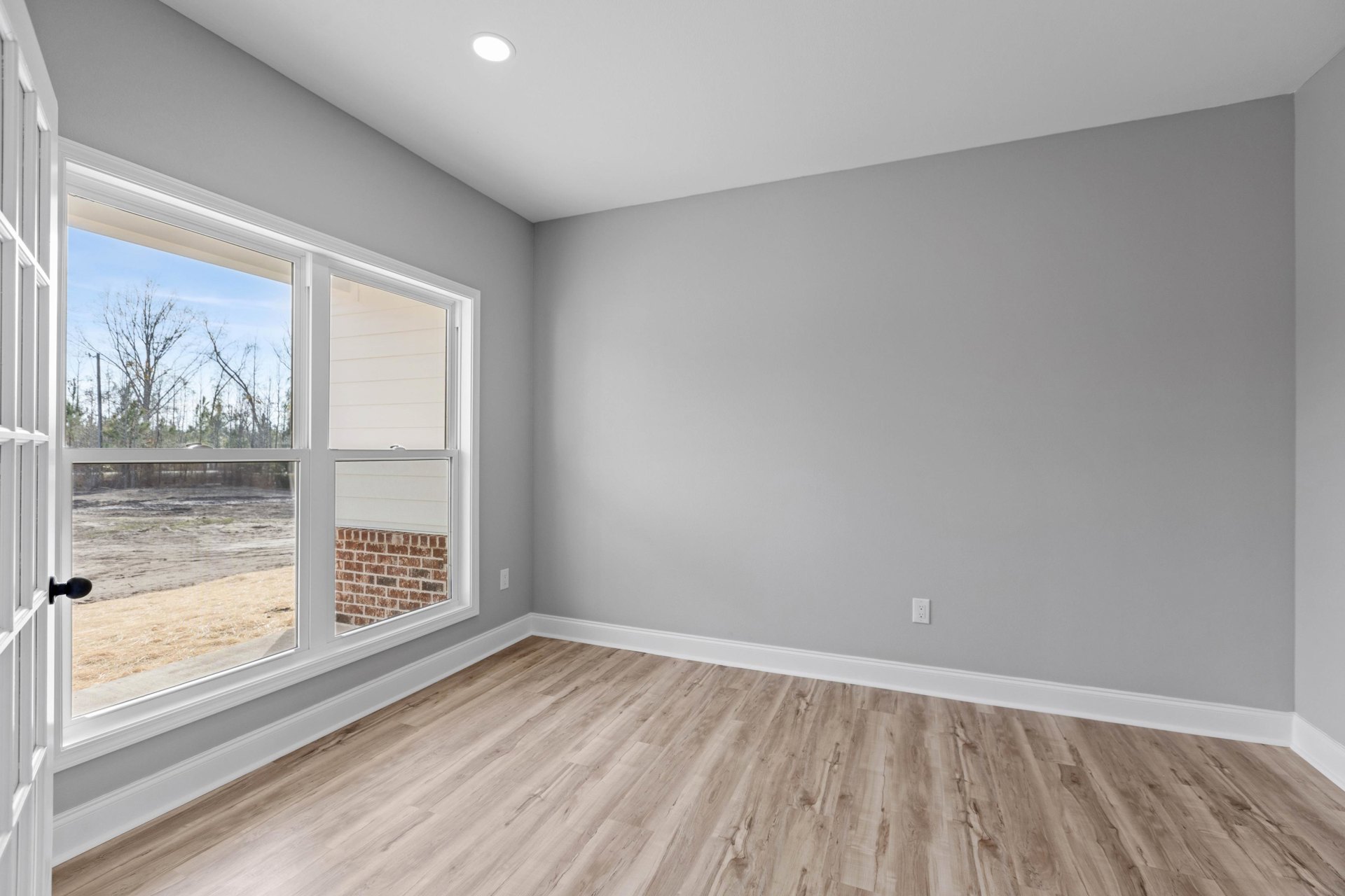 Wood flooring with white baseboards, large window overlooking leafless tree and dirt field, white ceiling featuring black stripe