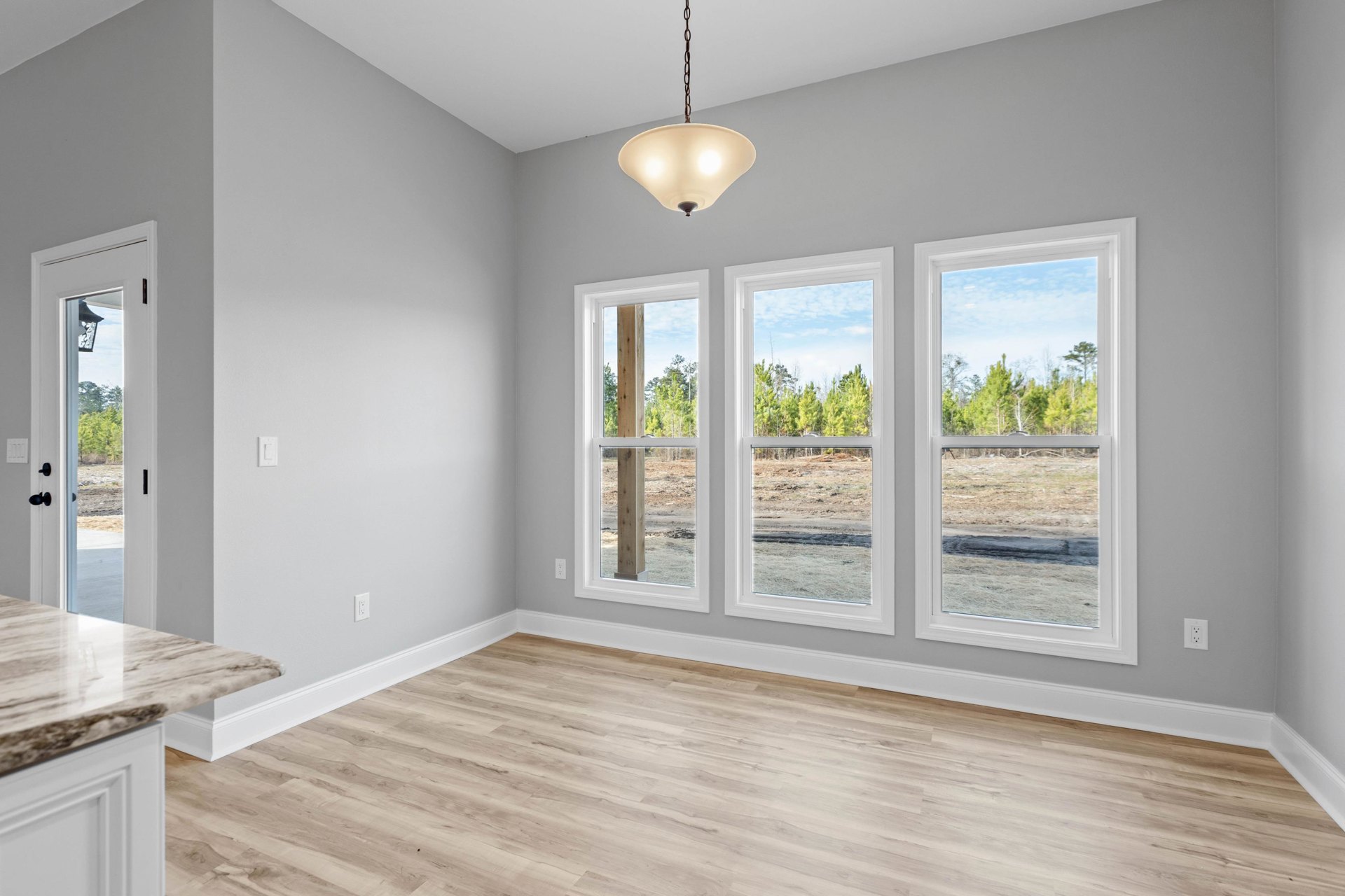 Sunlit room featuring large windows, warm wood flooring, white walls, ceiling-mounted light fixture, and marble countertop visible in the foreground.