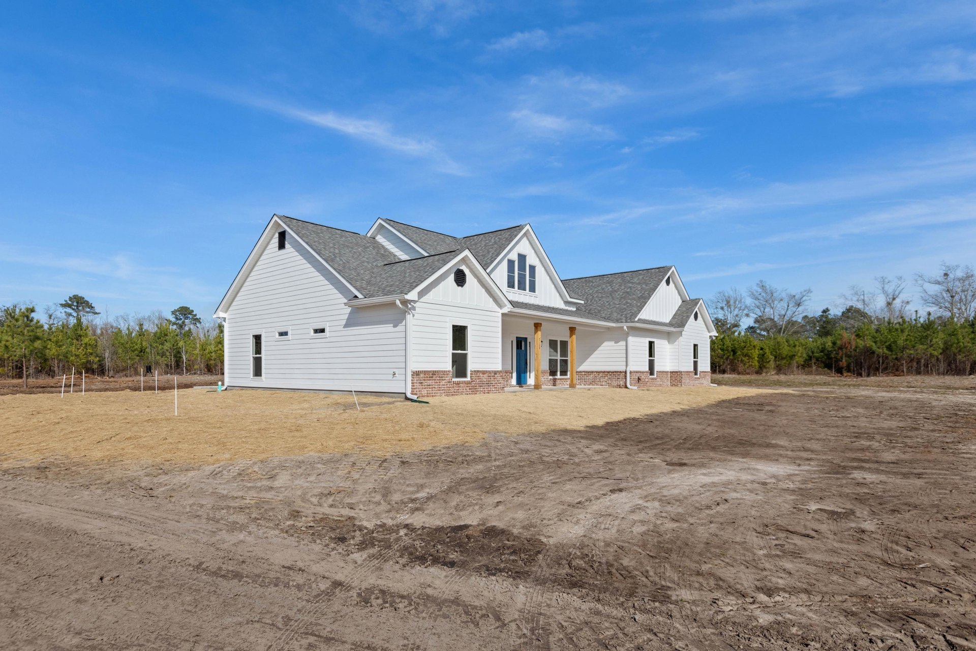 White house with brick accents and blue door, surrounded by dirt lot and scattered trees under clear blue sky
