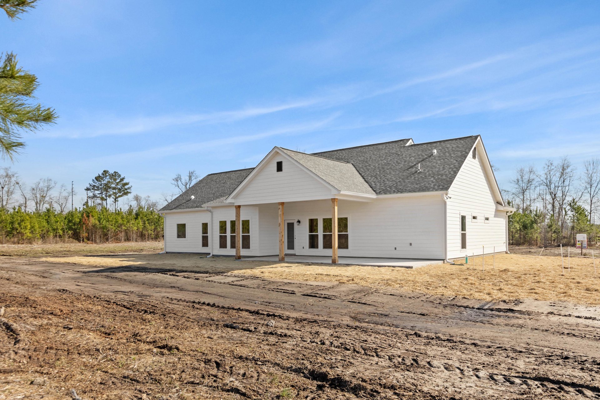 White siding house with covered porch, unfinished dirt yard, gray shingle roof, tree branch in foreground, partly cloudy sky