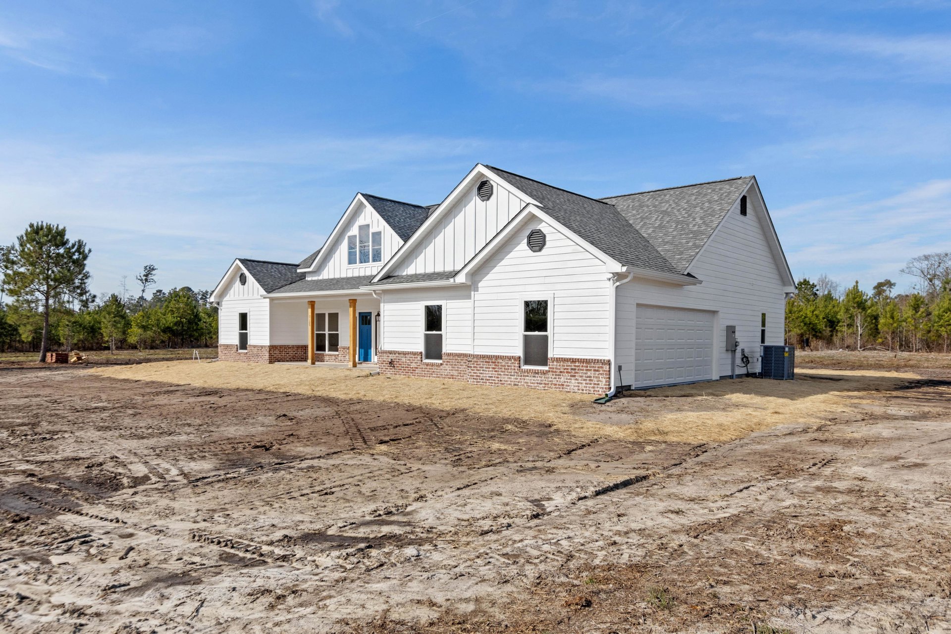 White custom home under construction with brick and wood siding, dirt lot in foreground, mature trees and blue sky in background