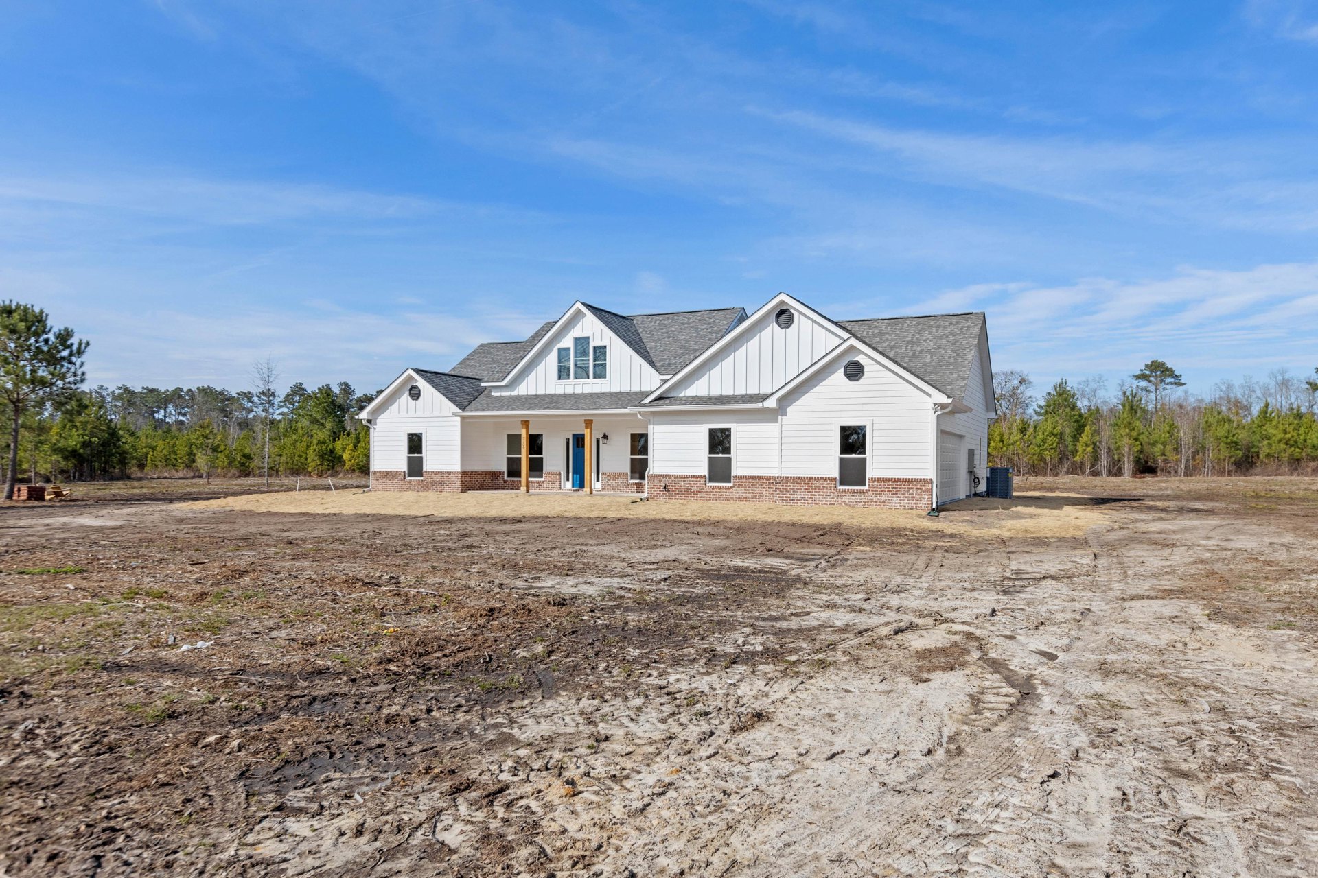 White house under construction with exposed framing, blue front door, dirt lot in foreground, and trees in background under a clear blue sky