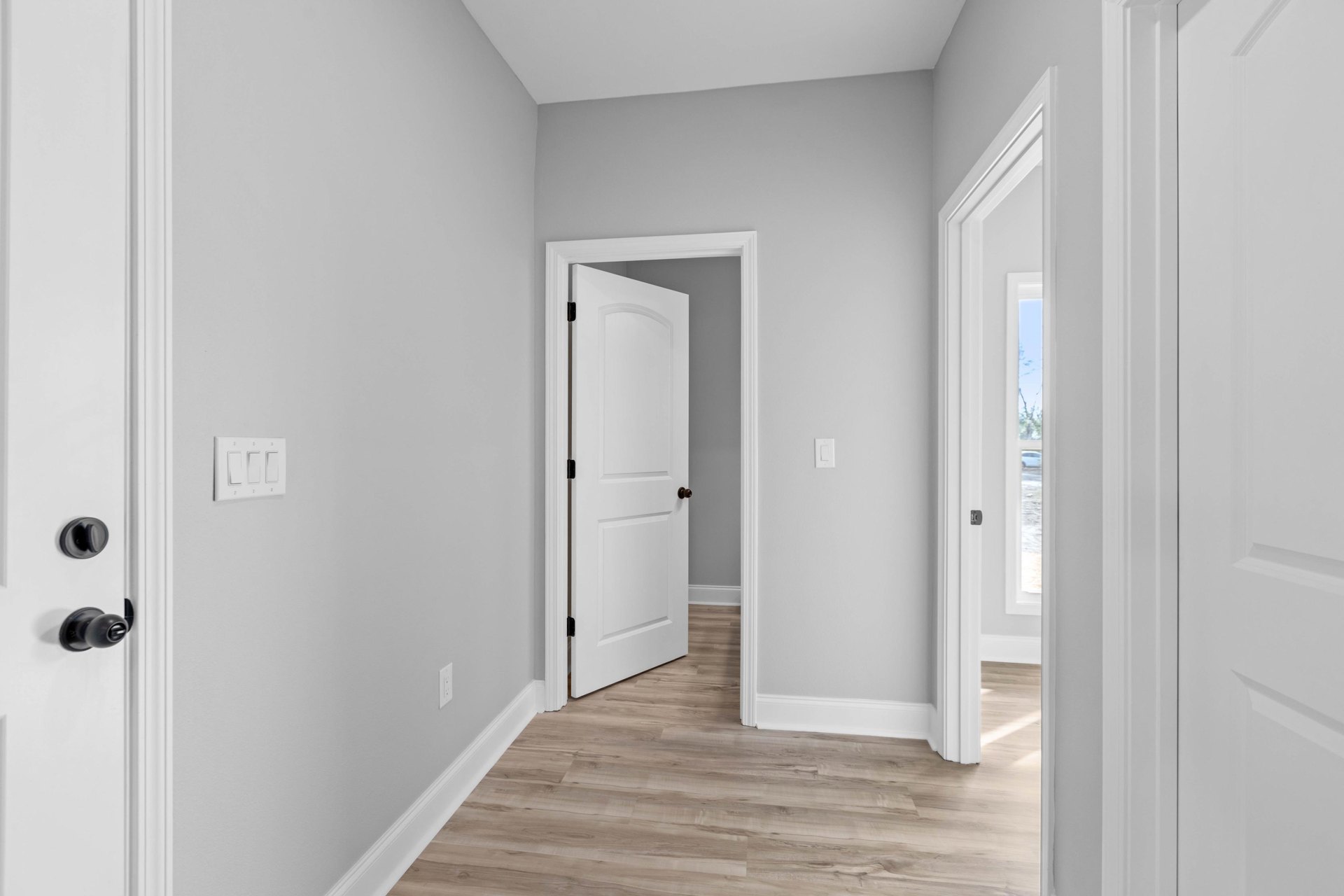 White hallway with wood flooring, white trim, and an open white door featuring a black handle; close-up details include a door knob and light switch on the wall.