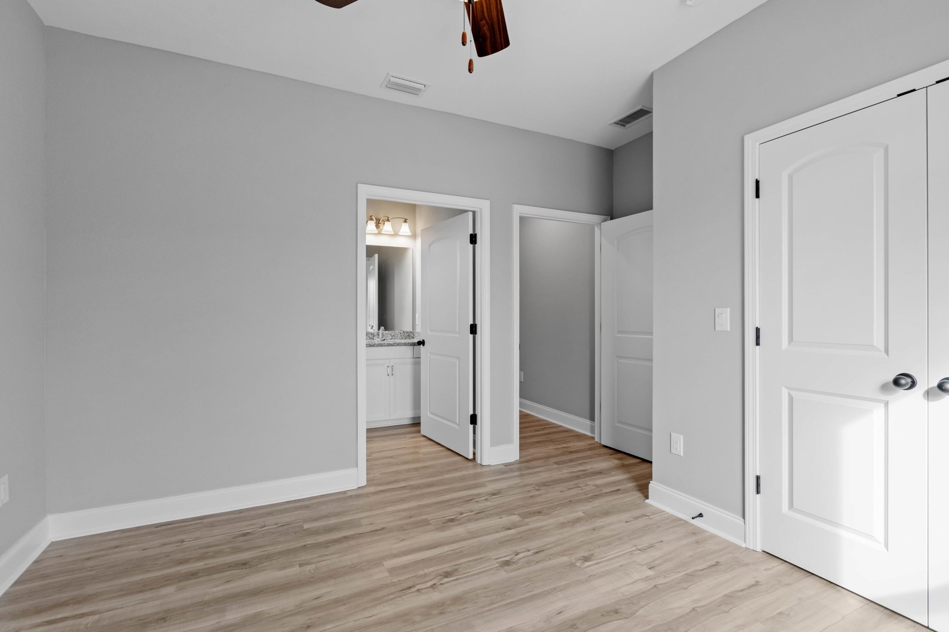 White paneled door with black knobs opening into a room featuring wood flooring, white walls, ceiling vent, and a ceiling fan with dark blades.