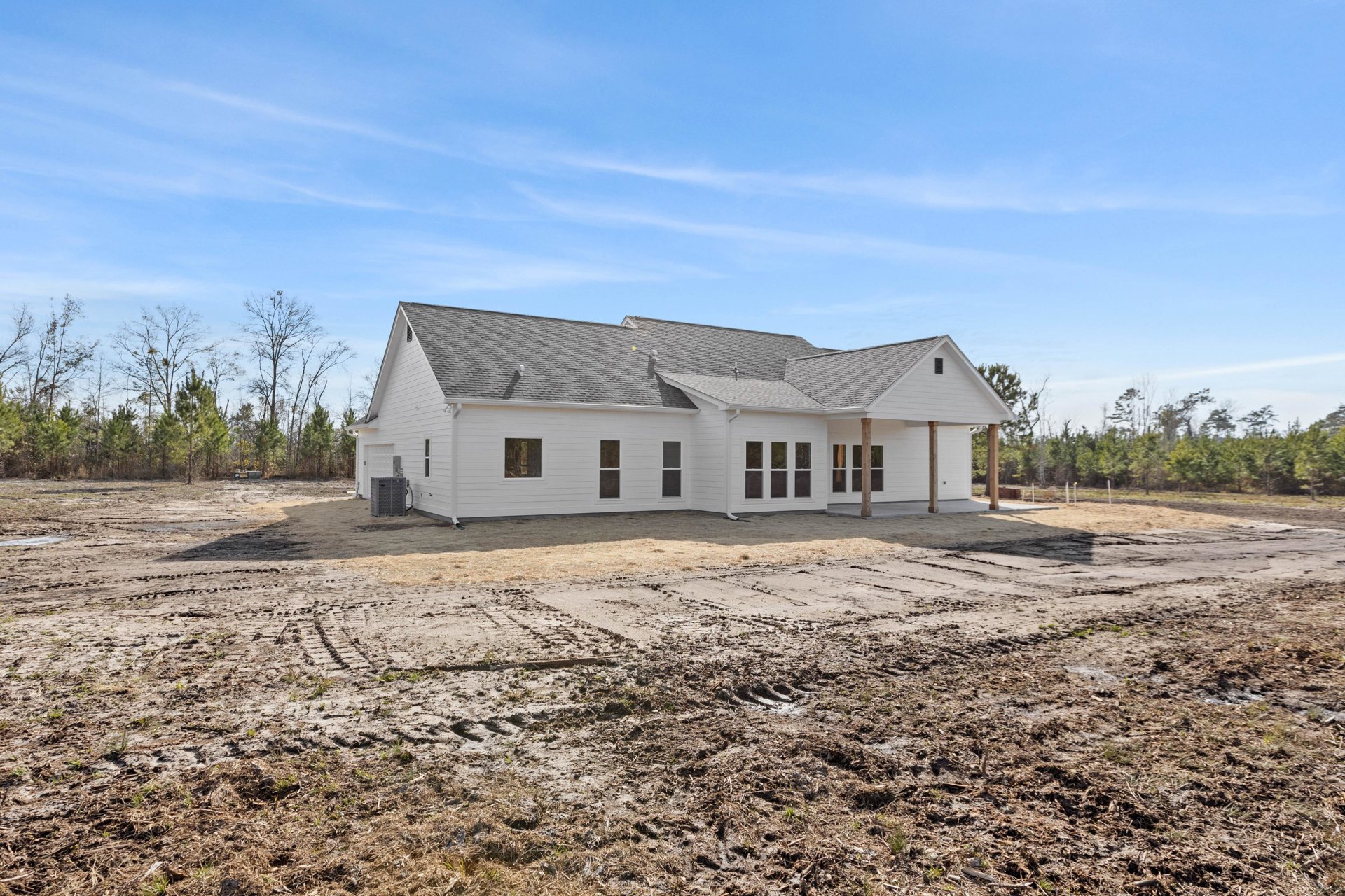 White house under construction with exposed dirt lot, scattered trees in background, unfinished windows, and rooftops beneath blue sky