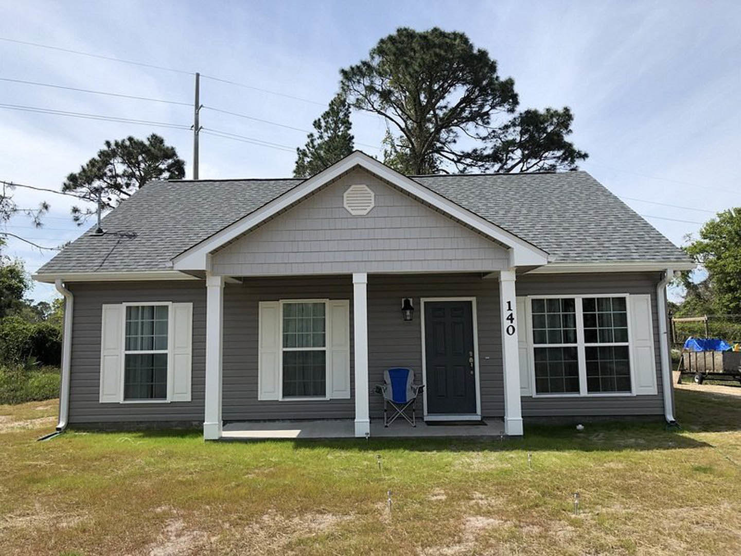 Gray house exterior with white trim, black front door, window with white shutters, blue and white folding chair on porch, green lawn, trees in background