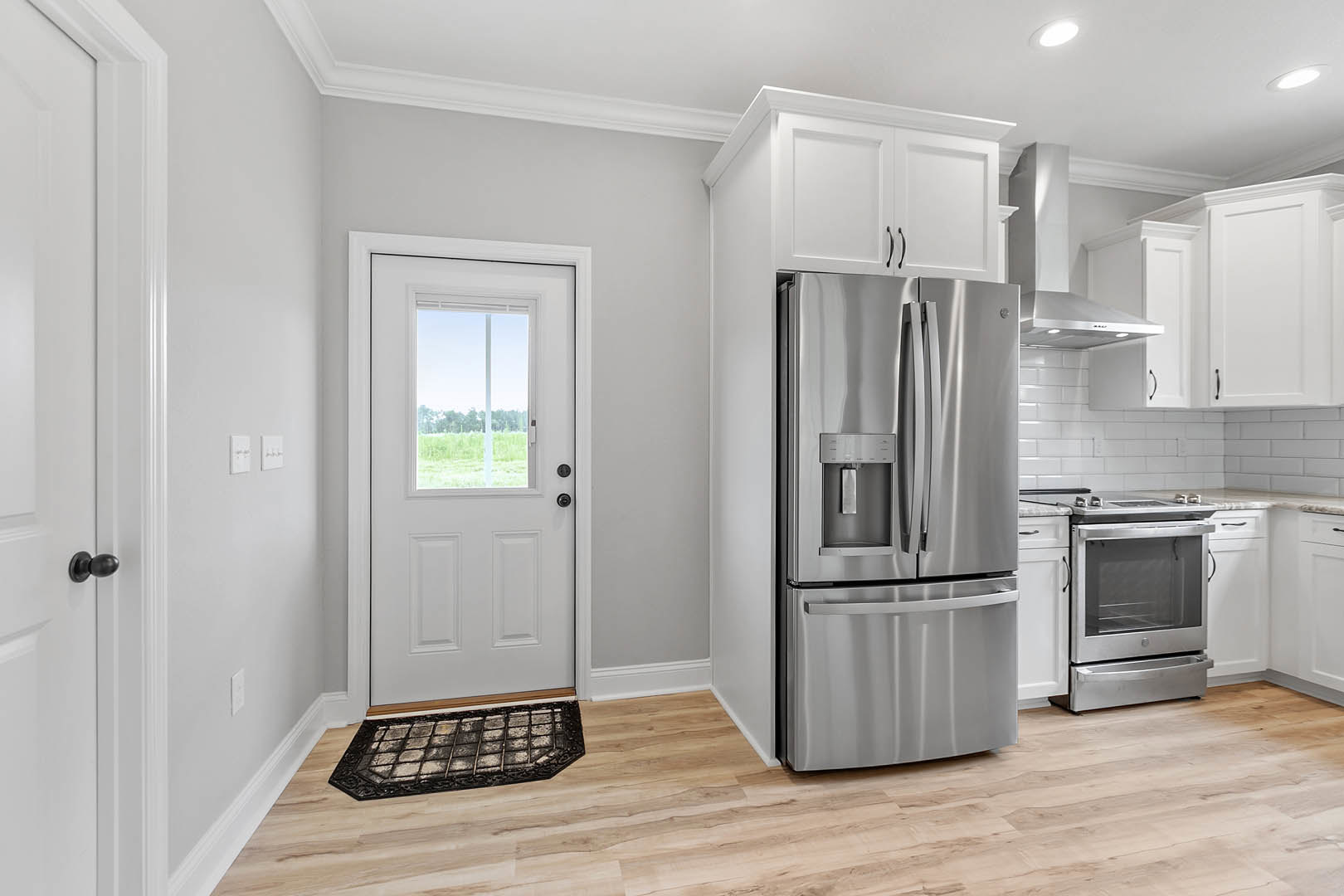 Stainless steel refrigerator with water dispenser beside white door with window, black door knob, white cabinetry, oven close-up, dirty wall grate, light countertops, wood flooring