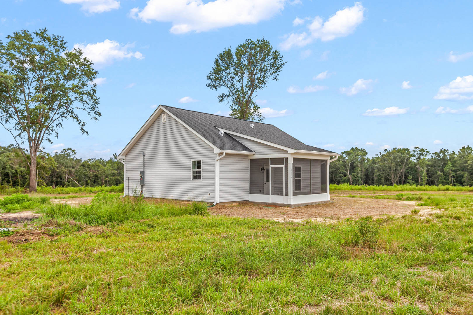 White farmhouse with covered front porch, surrounded by green grass field and mature leafy tree, under partly cloudy sky