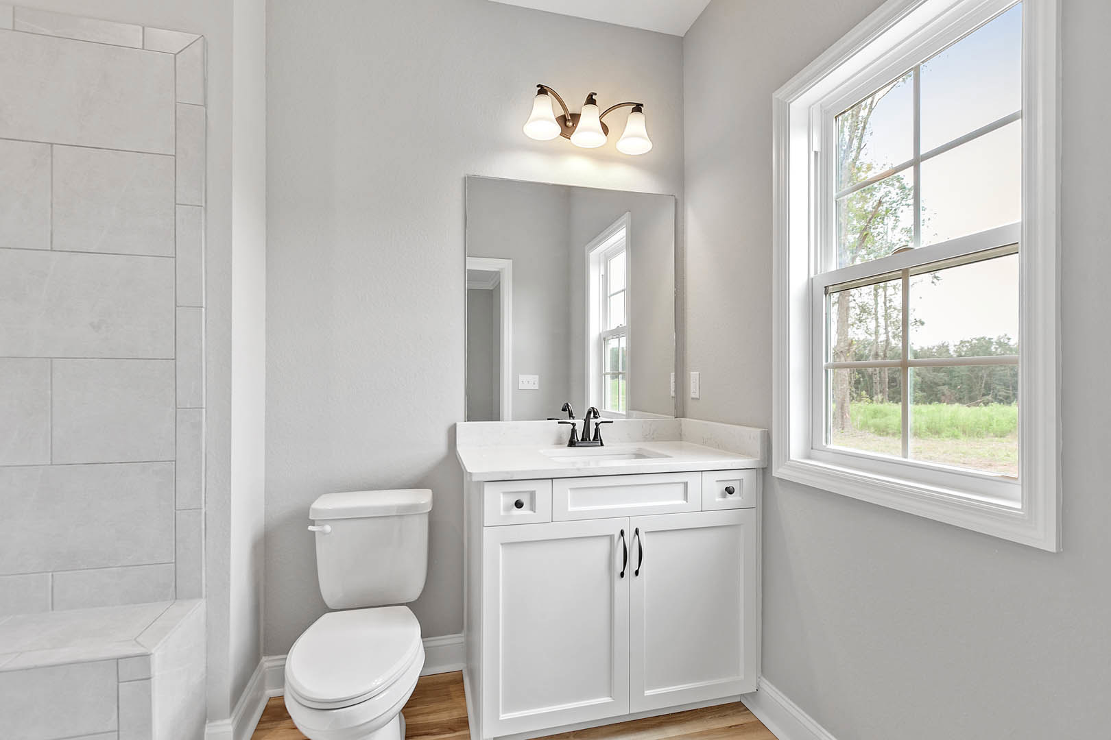 White tile bathroom featuring a white toilet with lid up, white sink with chrome tap, wall-mounted mirror, and window overlooking trees and grass.