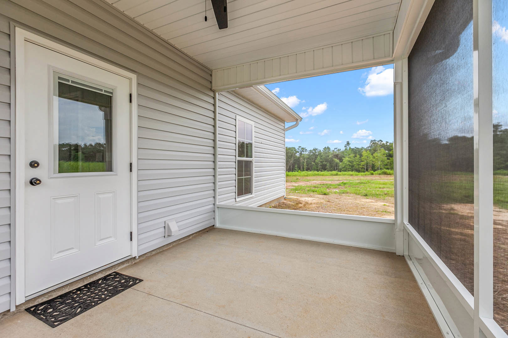 White front door with glass window and screen door opening onto covered porch, white-framed window to the side, textured floor with doormat, leafy trees visible through window.
