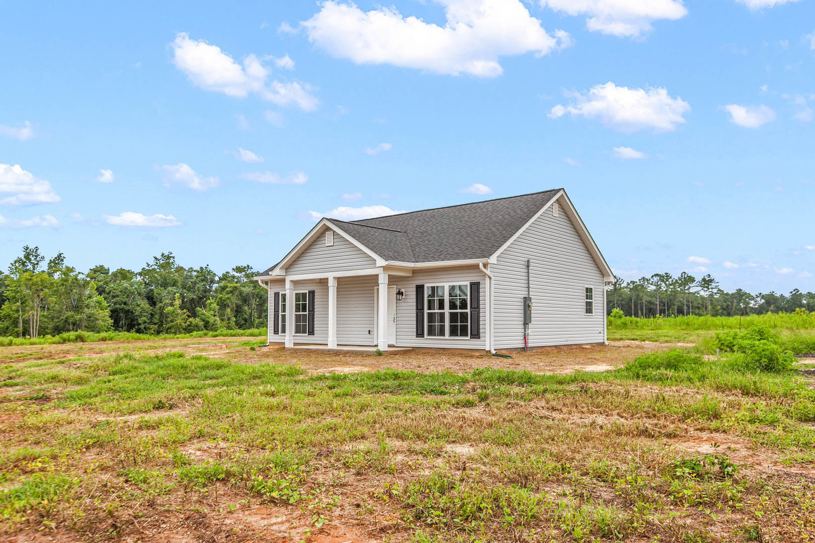 White siding house with covered front porch, large window, and gabled roof, set in a grassy field with trees in the background under a clear blue sky